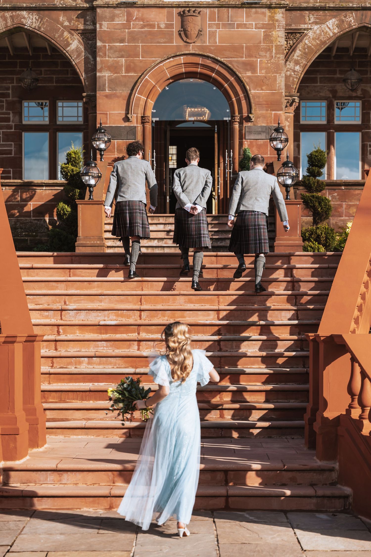 Wedding party members walk up red brick stairs at historic Boclair House entrance while bride waits below in flowing gown.