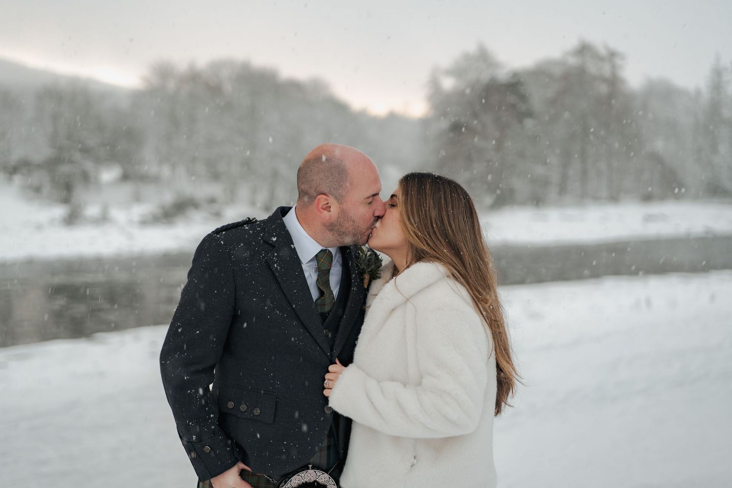 Couple sharing a kiss in a snowy winter landscape, man in kilt and woman in white fur coat.