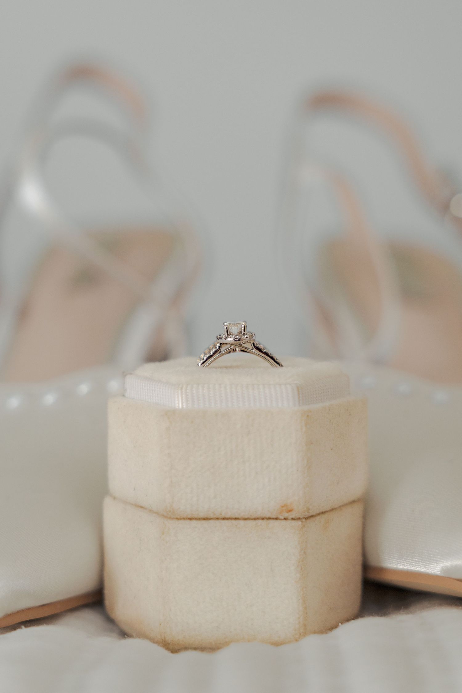 A delicate diamond engagement ring sits atop a cream-colored velvet ring box with elegant wedding shoes in the background.