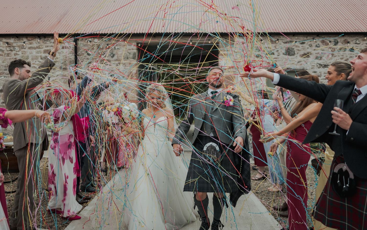 Bride and groom walk through colorful streamer confetti celebration outside rustic stone barn at Scottish wedding.