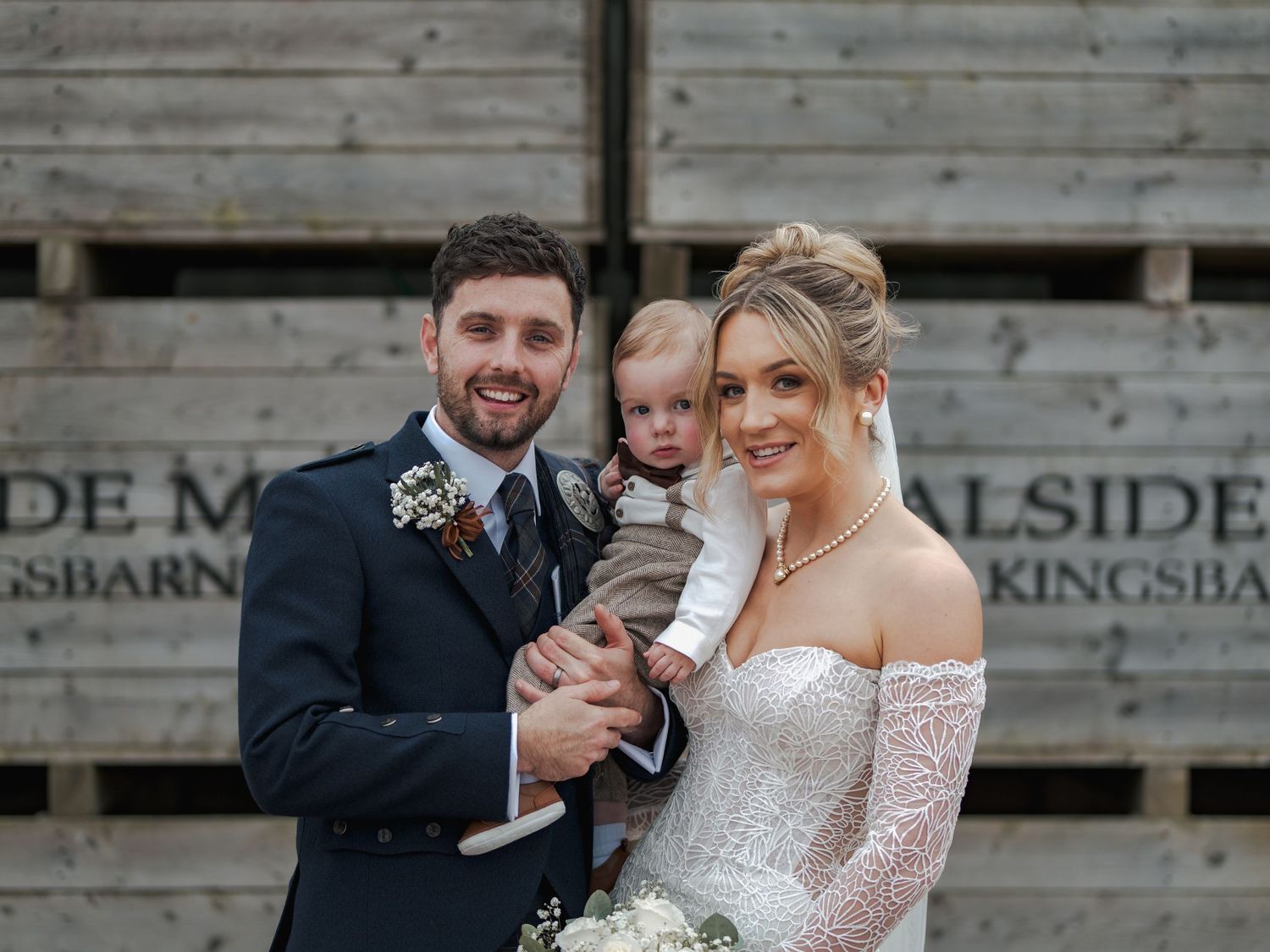 Newlywed couple shares joyful laughter during outdoor wedding photos against rustic wooden wall backdrop at Falside Mill.
