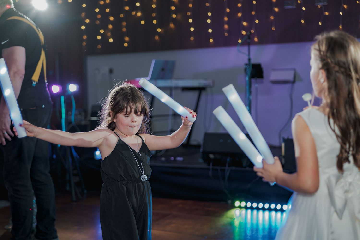 Children wave glow sticks while dancing at an indoor party with string lights and disco lighting in the background.