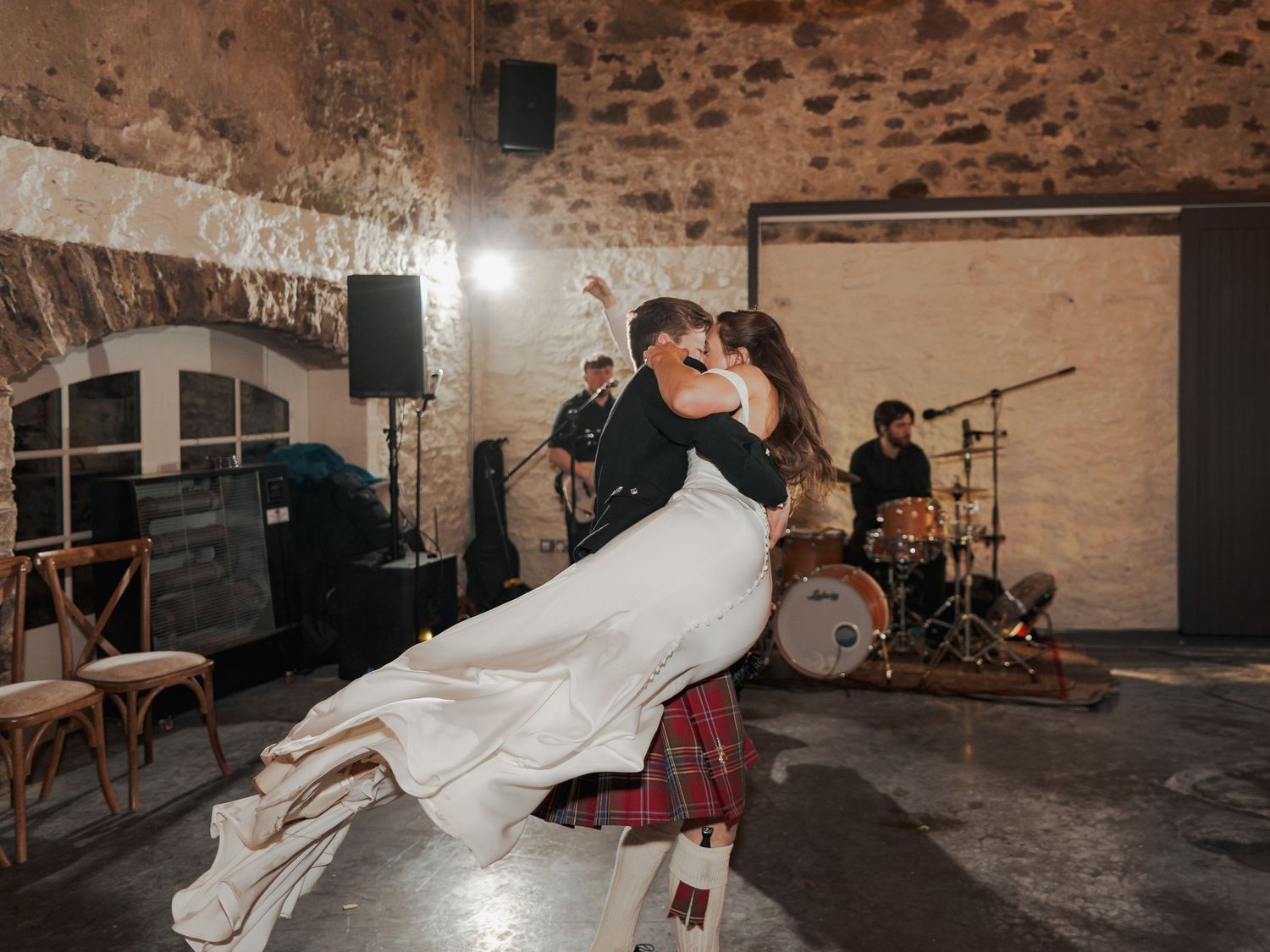 Couple shares romantic first dance dip in Scottish barn wedding venue with live band performing in background.