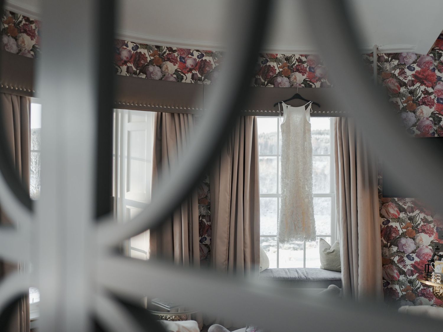 A wedding dress hanging in a floral-wallpapered room, viewed through a decorative mirror frame.