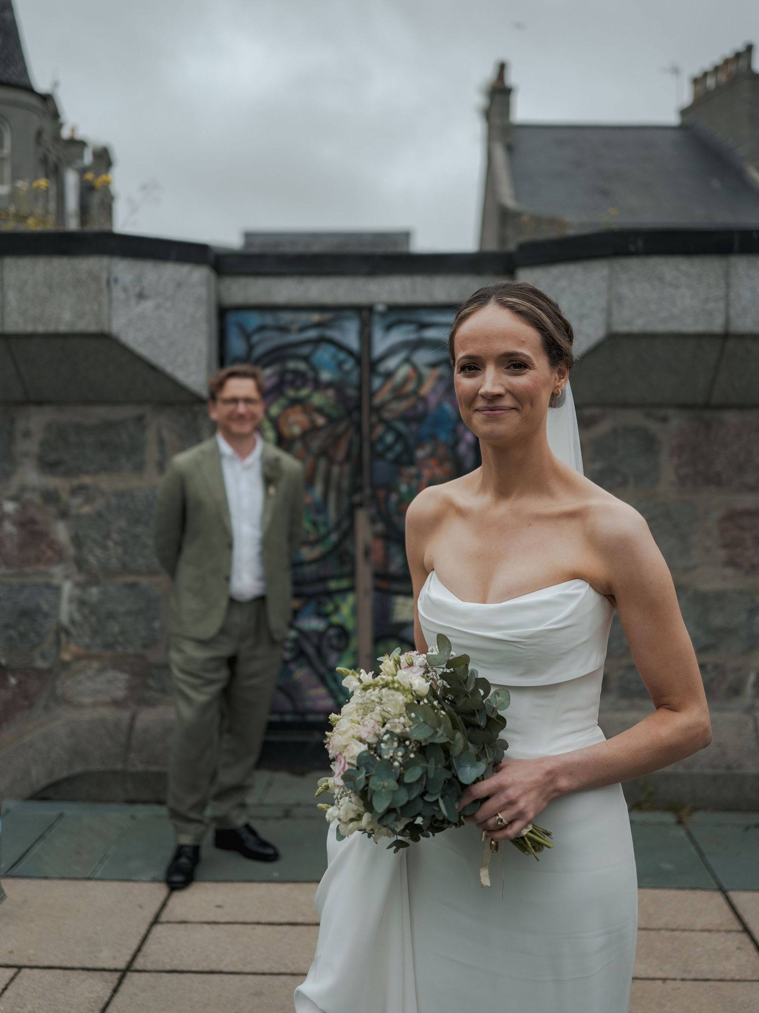 Bride in white strapless gown holding bouquet stands before stained glass church wall, groom in green suit behind her.