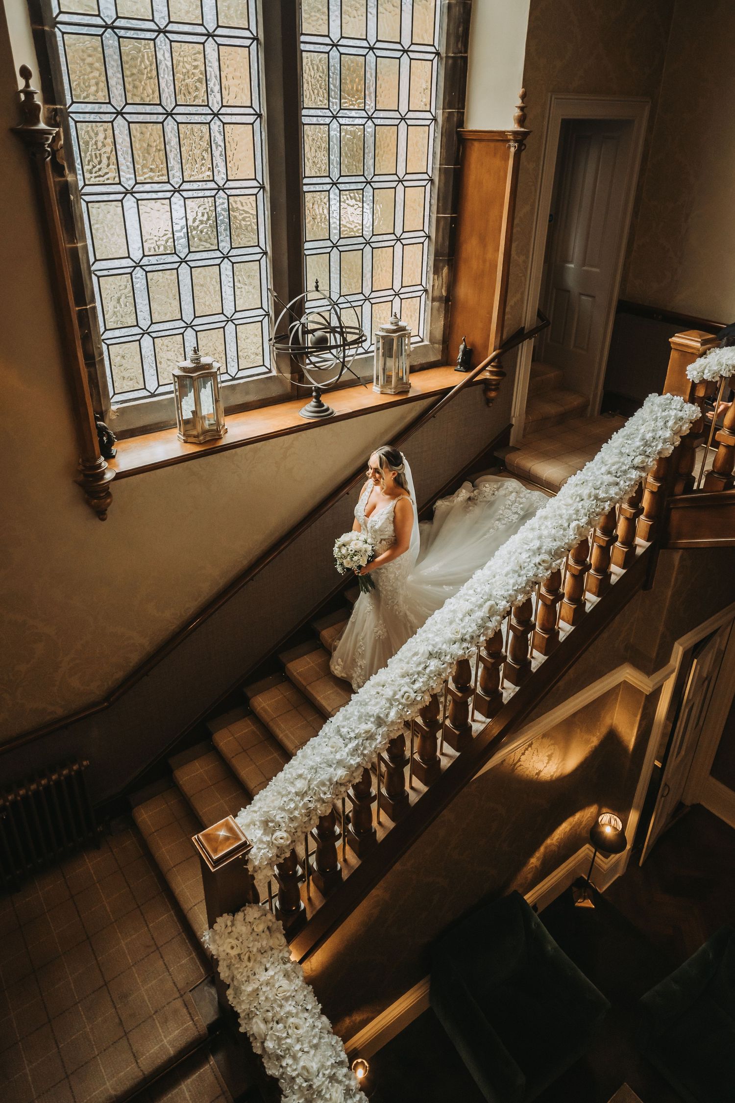 Bride descending ornate staircase decorated with white flowers in historic Cornhill Castle with stained glass window.