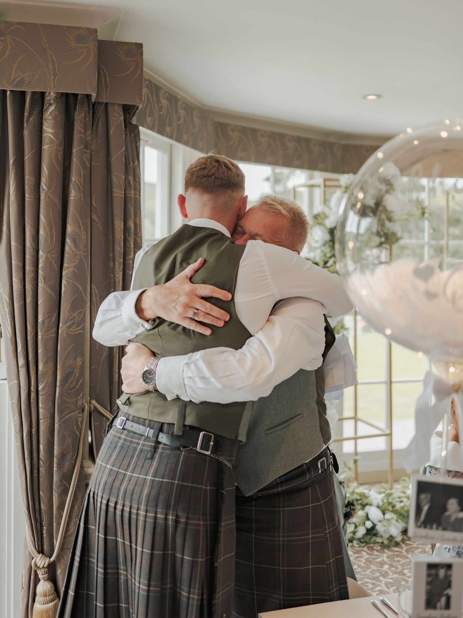 Two people in traditional Scottish kilts and formal attire embrace warmly at an indoor event with elegant decor.