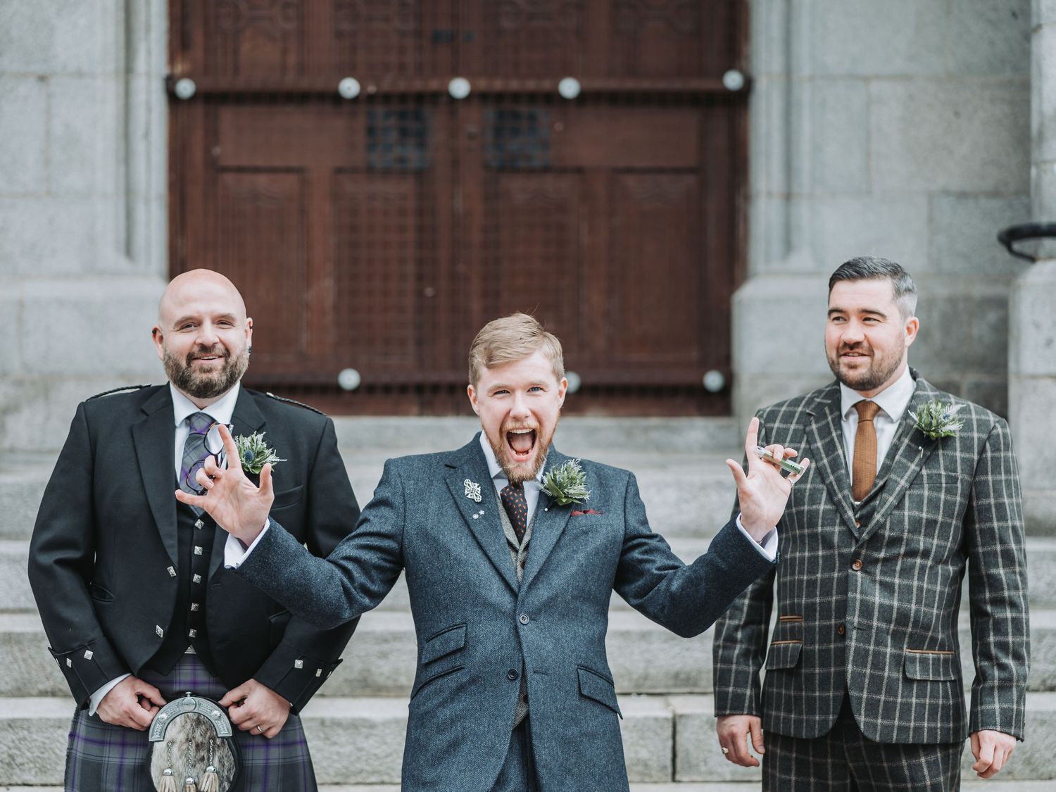 Three groomsmen in formal attire laugh together outside church doors at Marischal College.