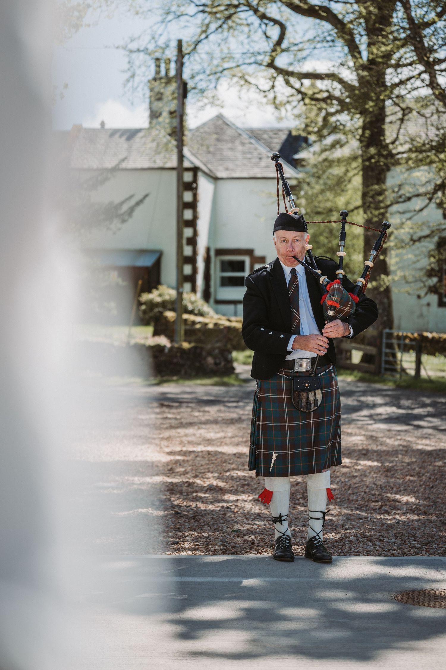 A bagpiper in traditional Scottish attire performs outdoors on a sunny day near Kinclune House.