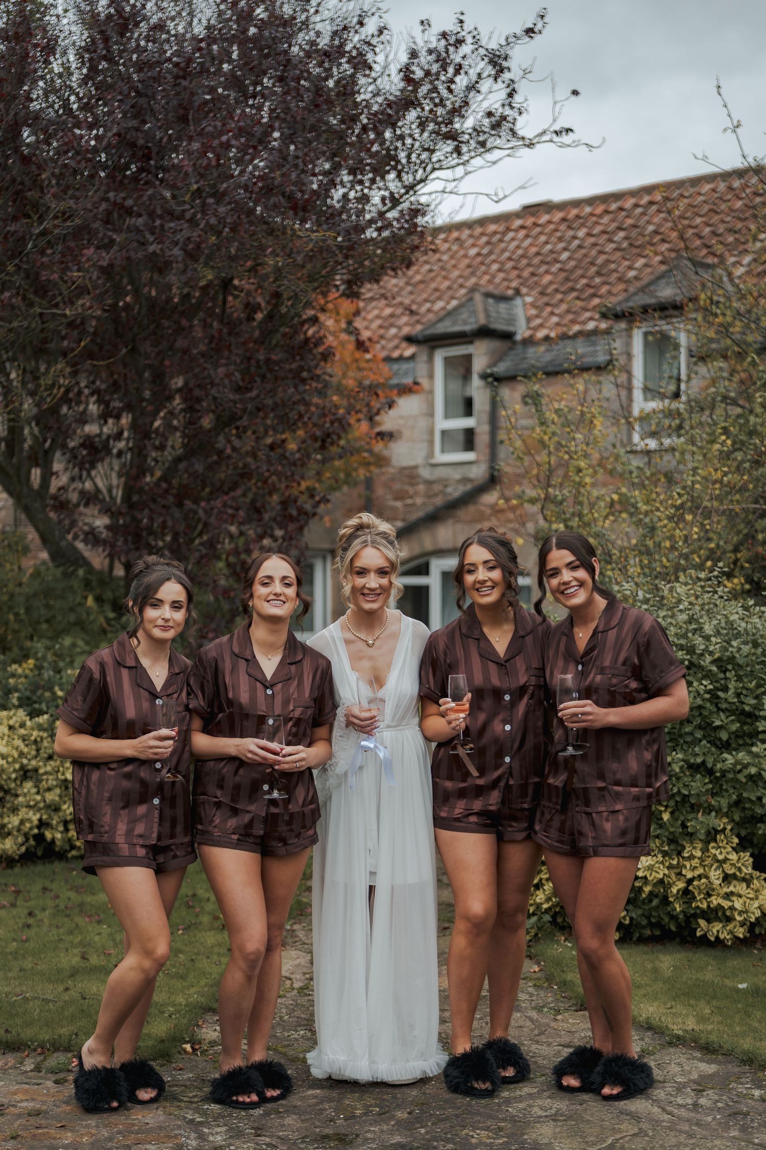 A bridal party poses in a garden, with four attendants wearing matching brown robes and the bride in a flowing blue gown.