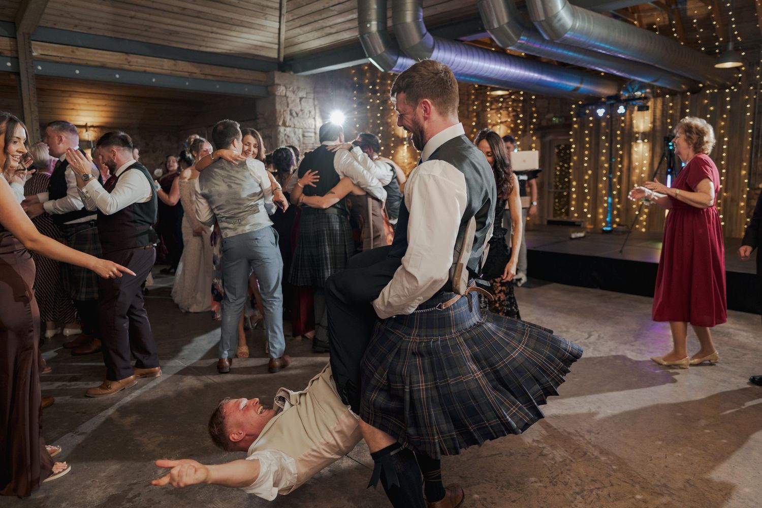 Partygoers dancing at Falside Mill beneath warm string lights in a rustic indoor venue.