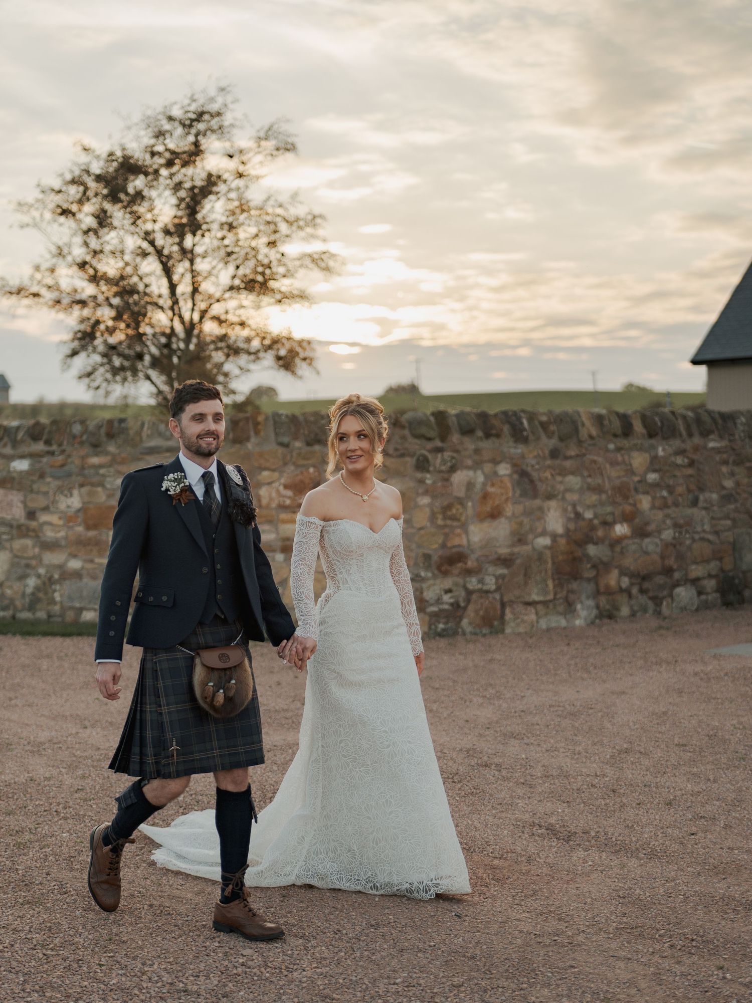 A romantic outdoor wedding photoshoot at Falside Mill against a stone wall at sunset with traditional Scottish attire.