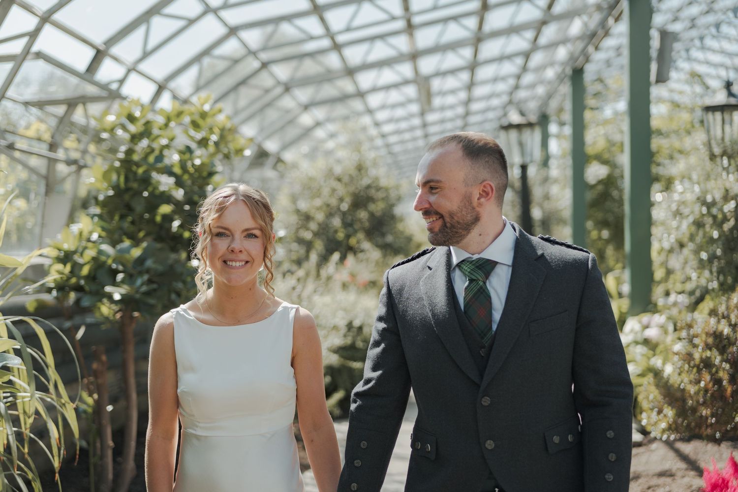 A happy couple shares a joyful moment together in a sunlit David Welsh Winter Gardens surrounded by lush greenery and glass walls.