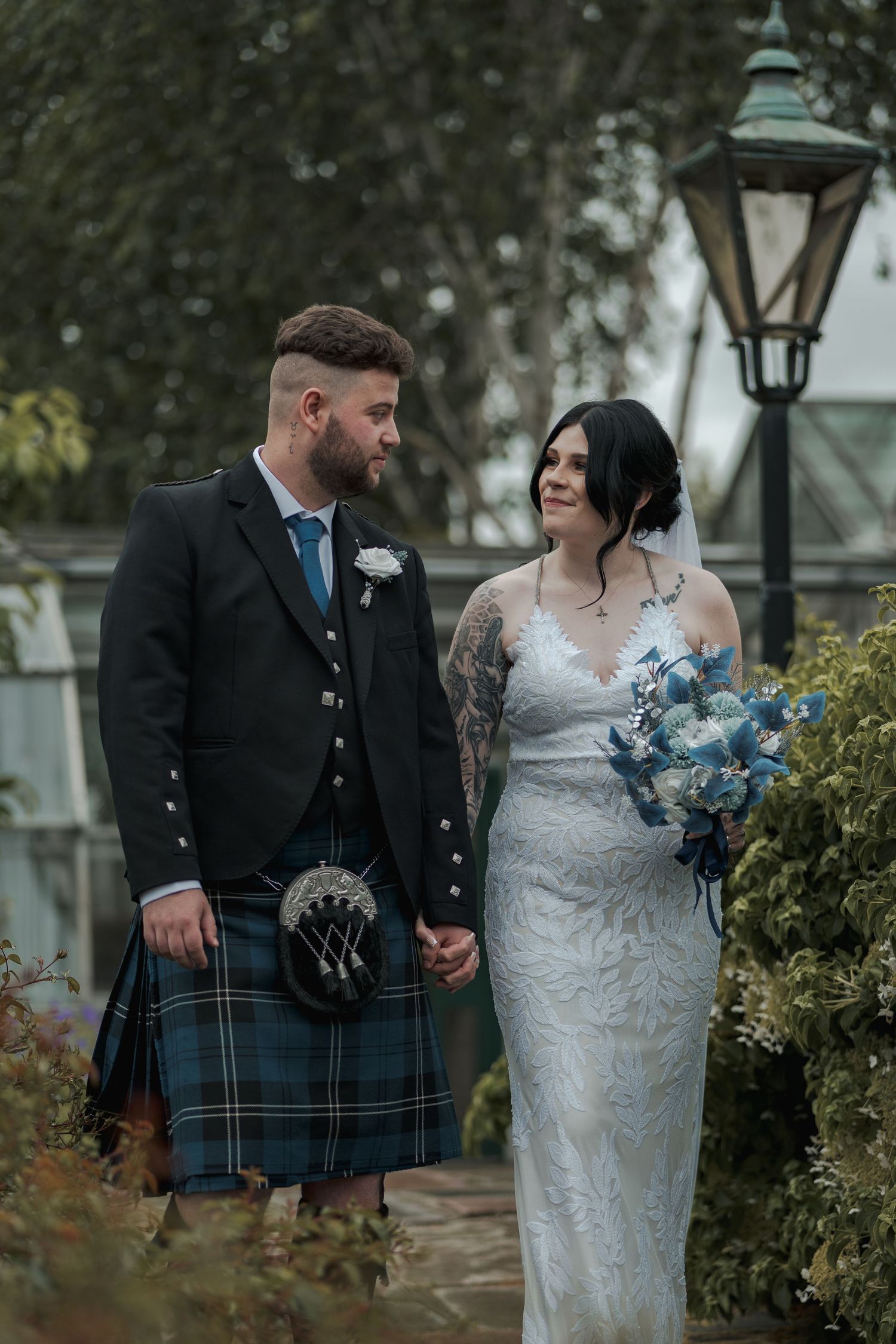 A couple in wedding attire, groom in Scottish kilt, bride in lace gown holding blue bouquet, posing in garden.
