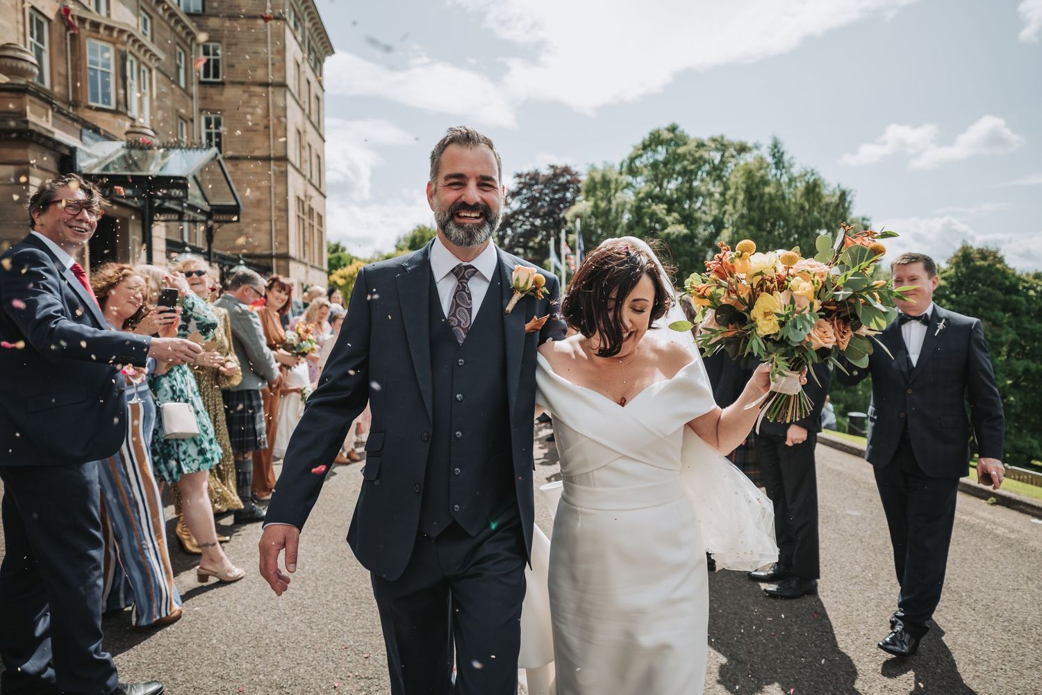 Wedding couple walks down steps of Dunblane Hydro with guests celebrating on both sides as bride holds colorful yellow bouquet.