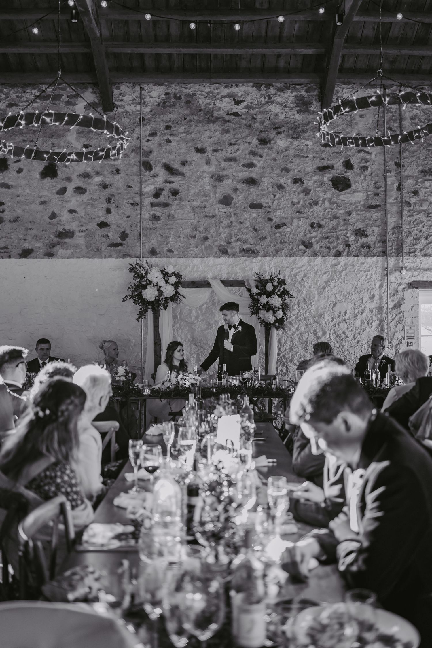 Black and white wedding reception photo showing guests dining at tables with elegant floral centerpieces in at rustic stone venue Kinclune.