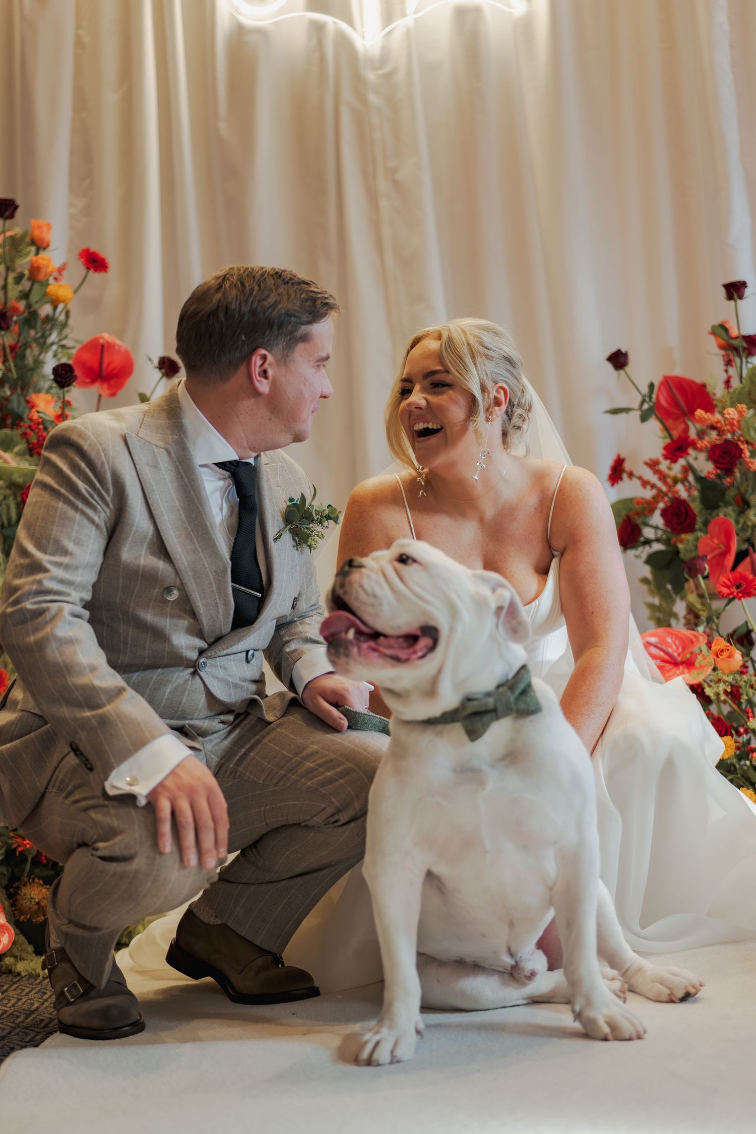 A white bulldog sits with a couple in formal attire against a backdrop of colorful flowers at the Sandman Signature, Aberdeen
