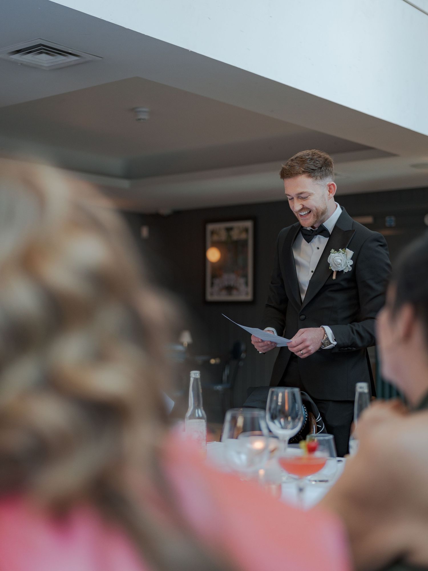 A person in a black suit reads from a paper during a formal dinner reception with wine glasses on tables.