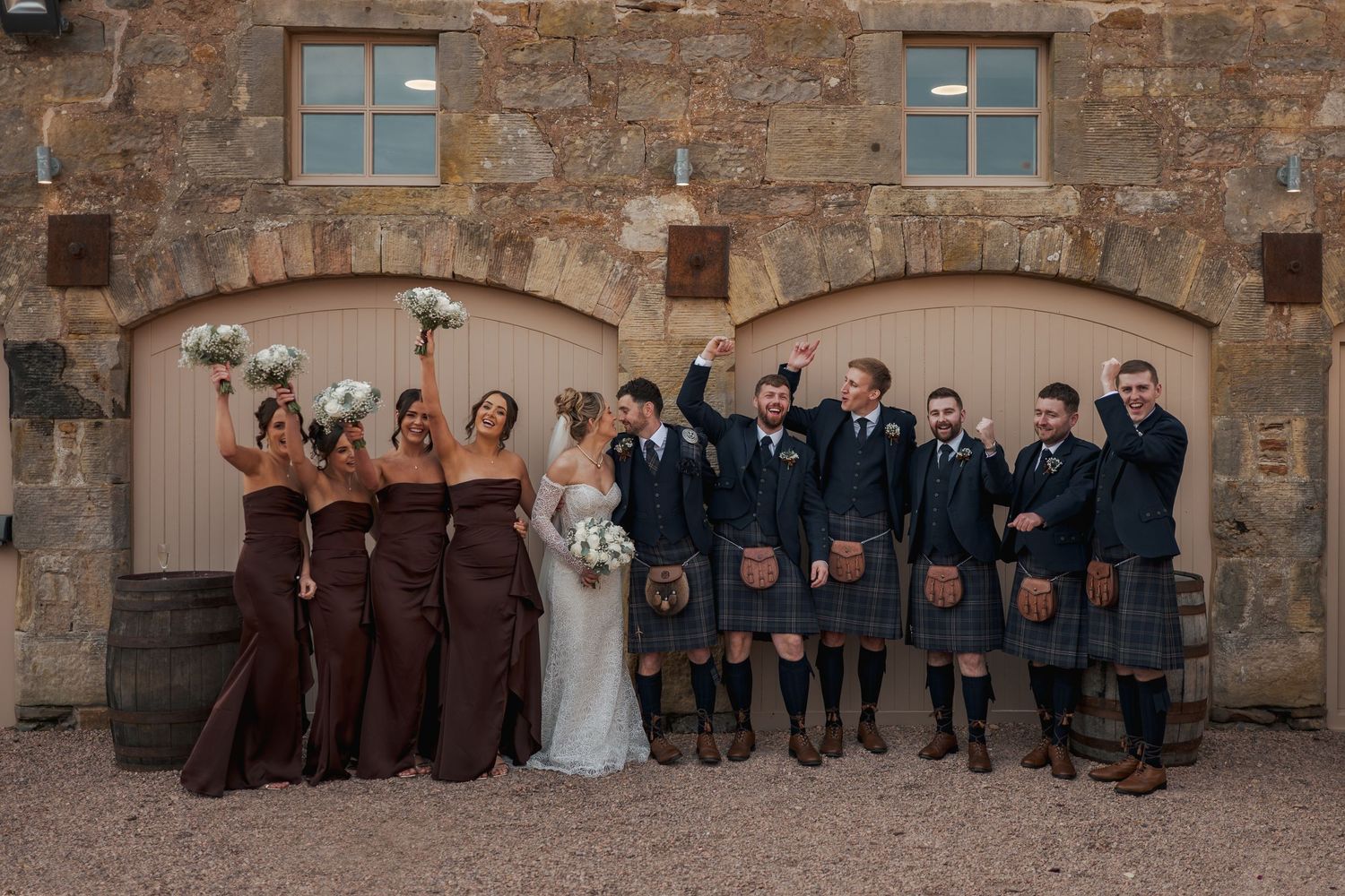 Wedding party in brown dresses and traditional kilts poses against rustic stone wall with arched doorways at Falside Mill.