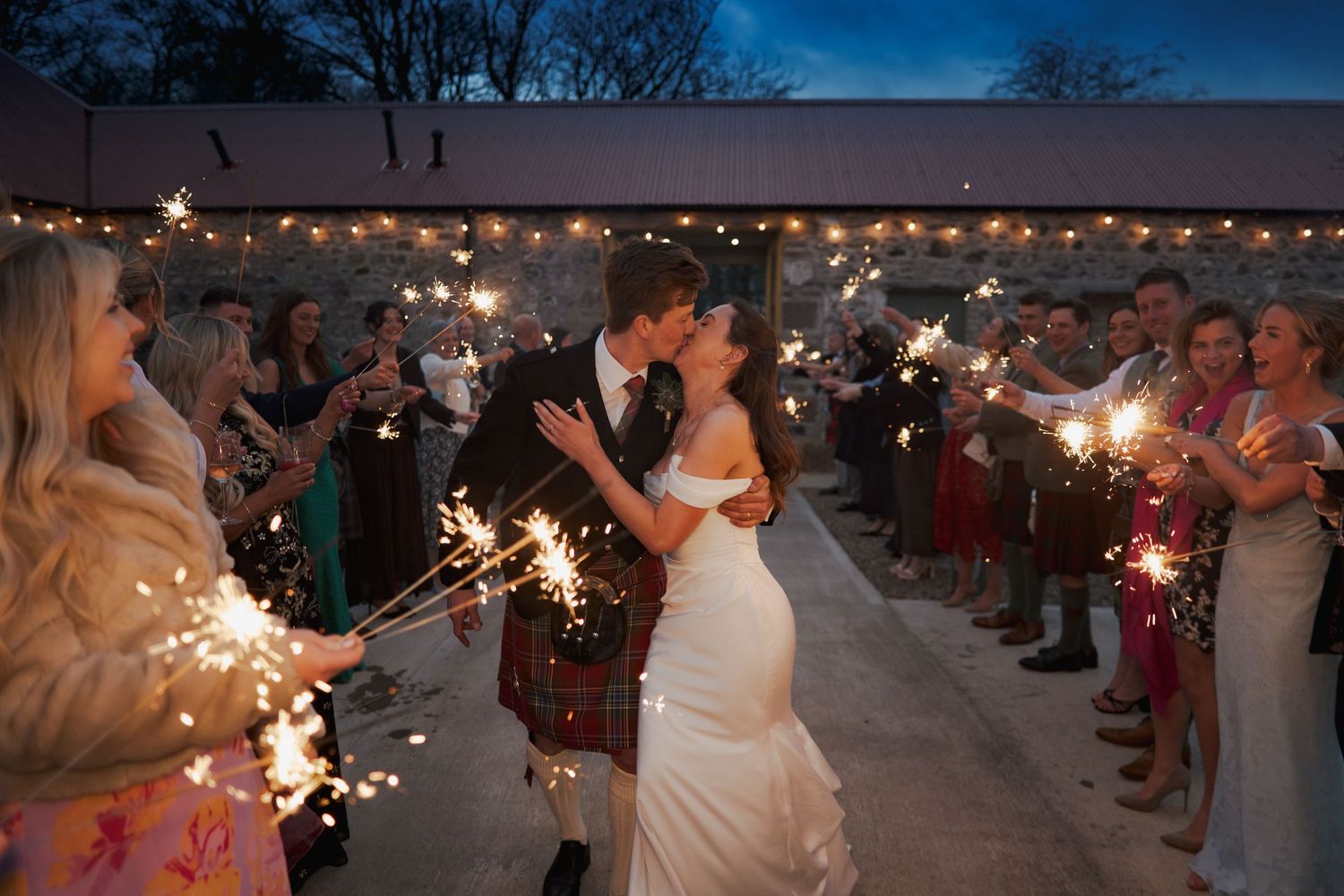 Bride and groom kissing during sparkler exit at twilight wedding celebration with guests holding sparklers.