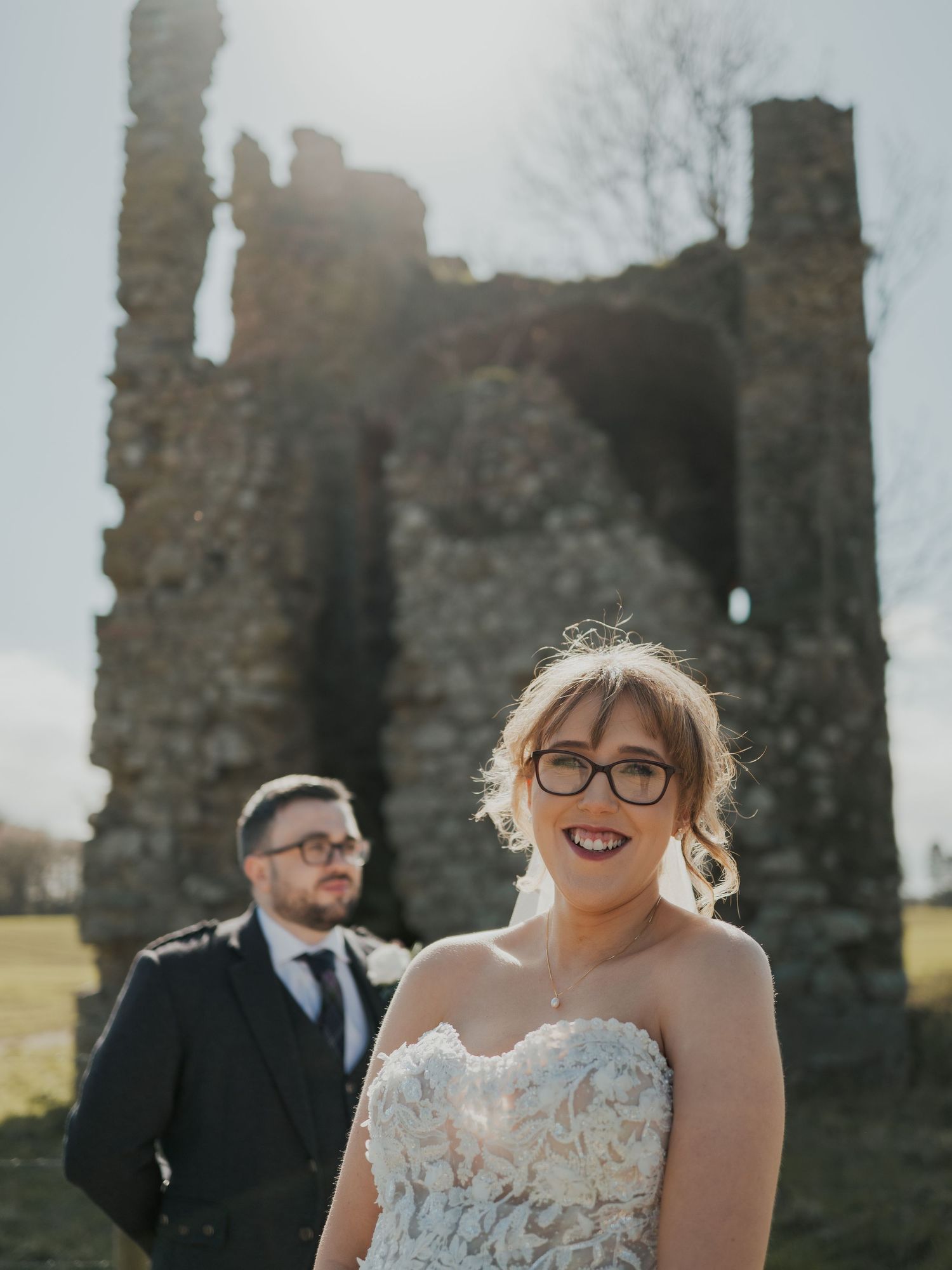 Bride in lace gown and groom in kilt pose before ancient stone ruins on a sunny wedding day.