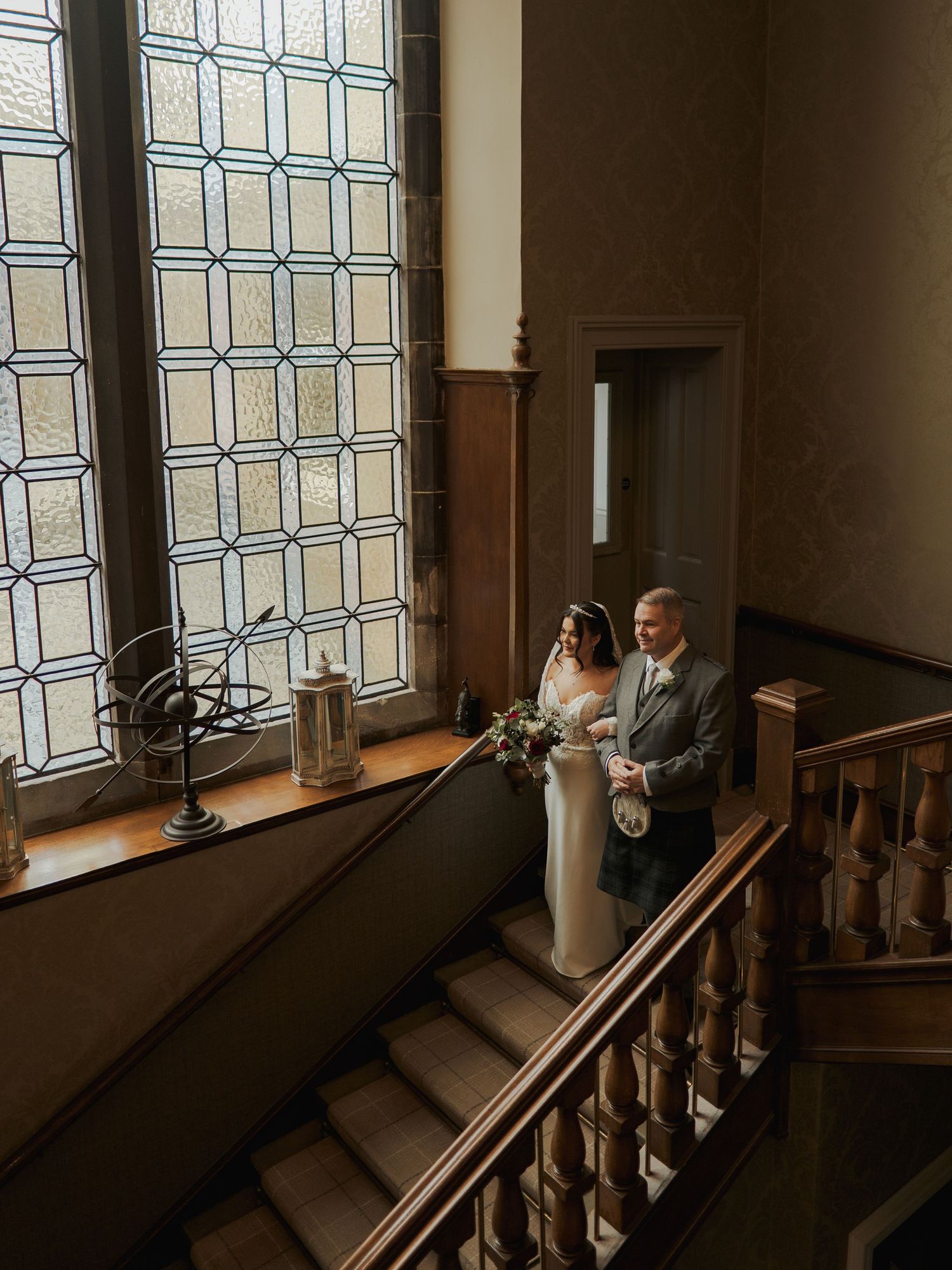 Bride and groom descend elegant staircase beside stained glass window in historic wedding venue.
