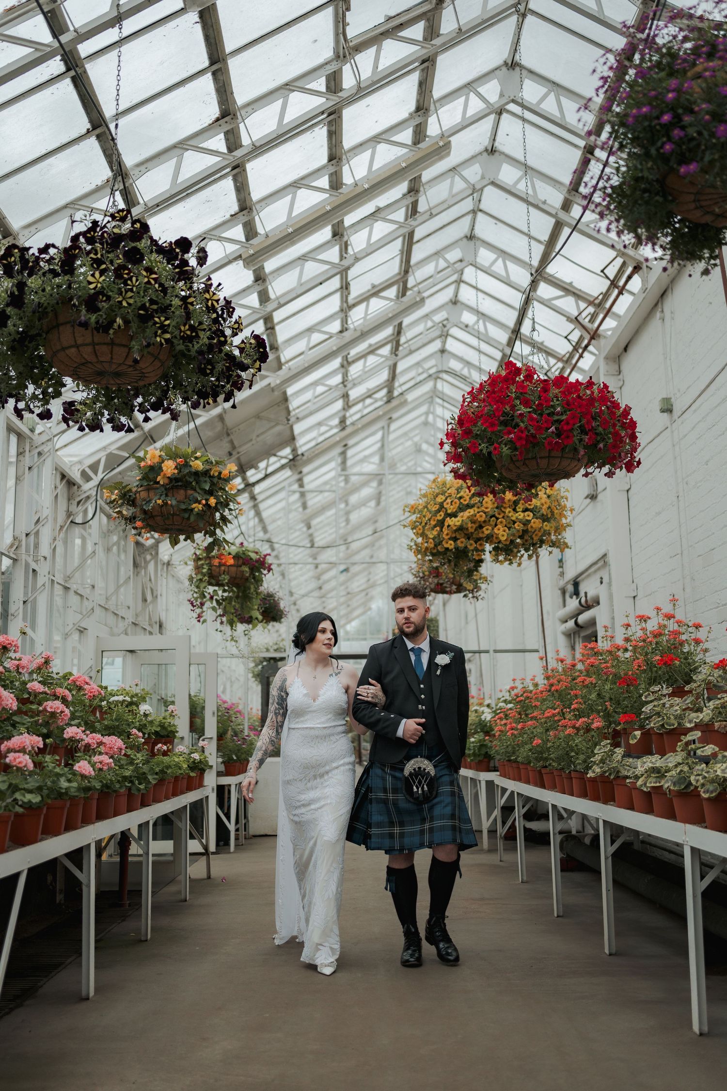 Bride and groom walk through a floral greenhouse, groom wearing a tartan kilt, surrounded by hanging flower baskets.