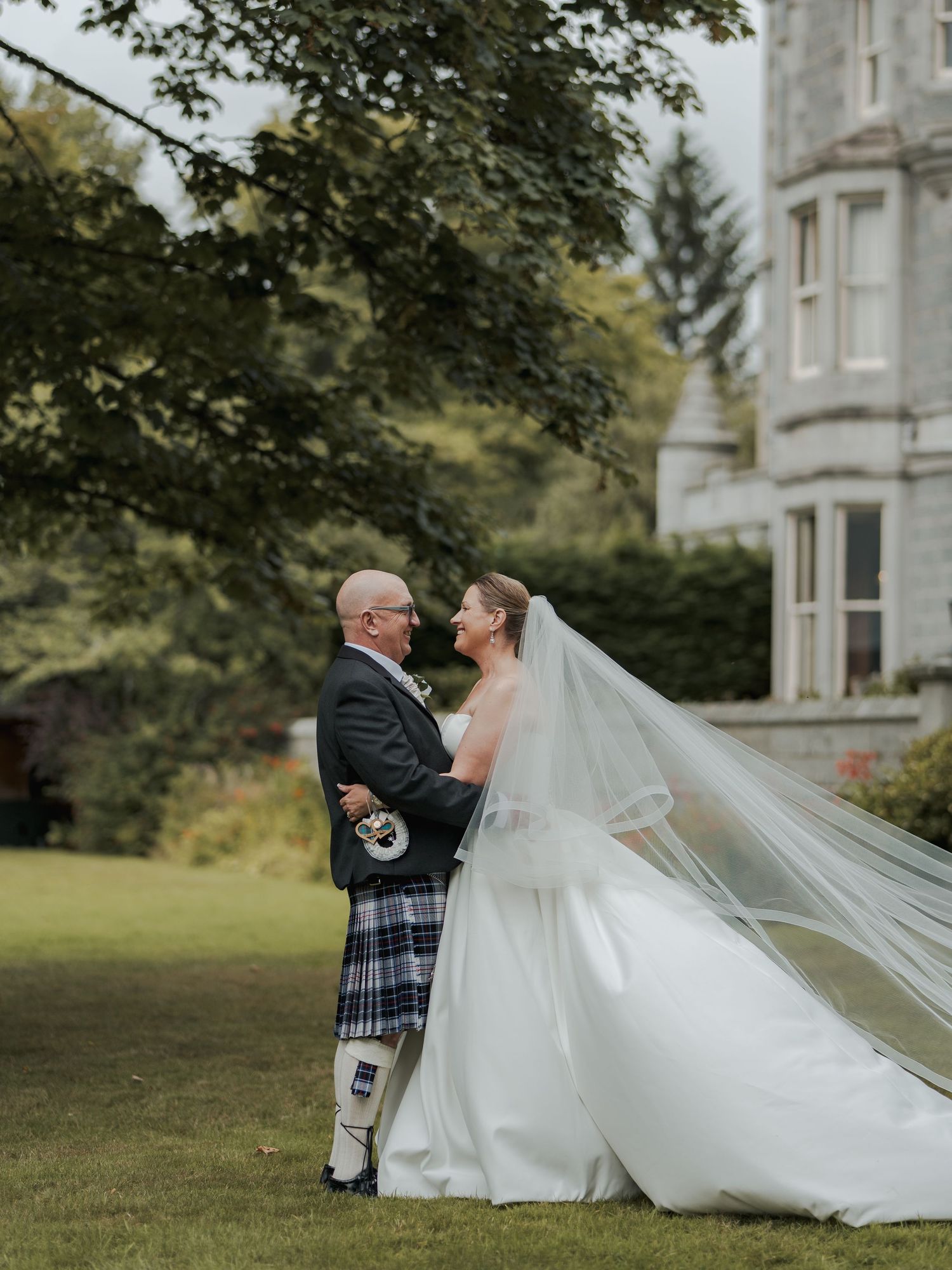 Bride and groom embrace on castle grounds, her veil flowing in the wind, groom wearing traditional Scottish tartan kilt.