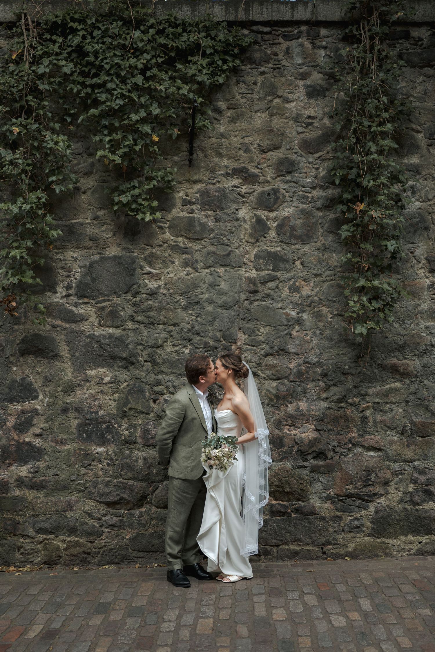 Bride and groom share a romantic kiss against an ivy-covered stone wall, bride holding white floral bouquet.