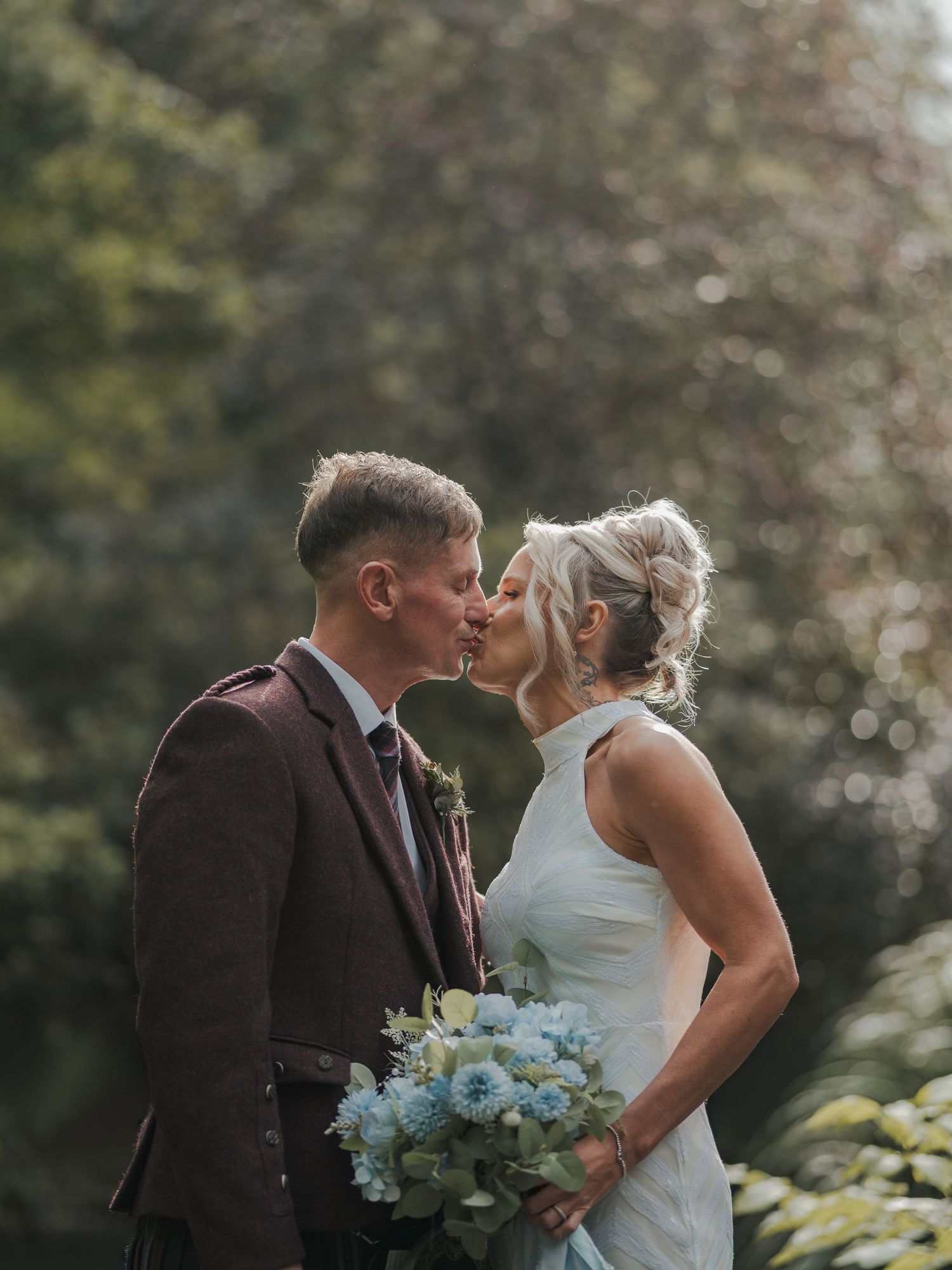 Bride and groom share a kiss outdoors, she holds blue bouquet, he wears dark suit with boutonniere.