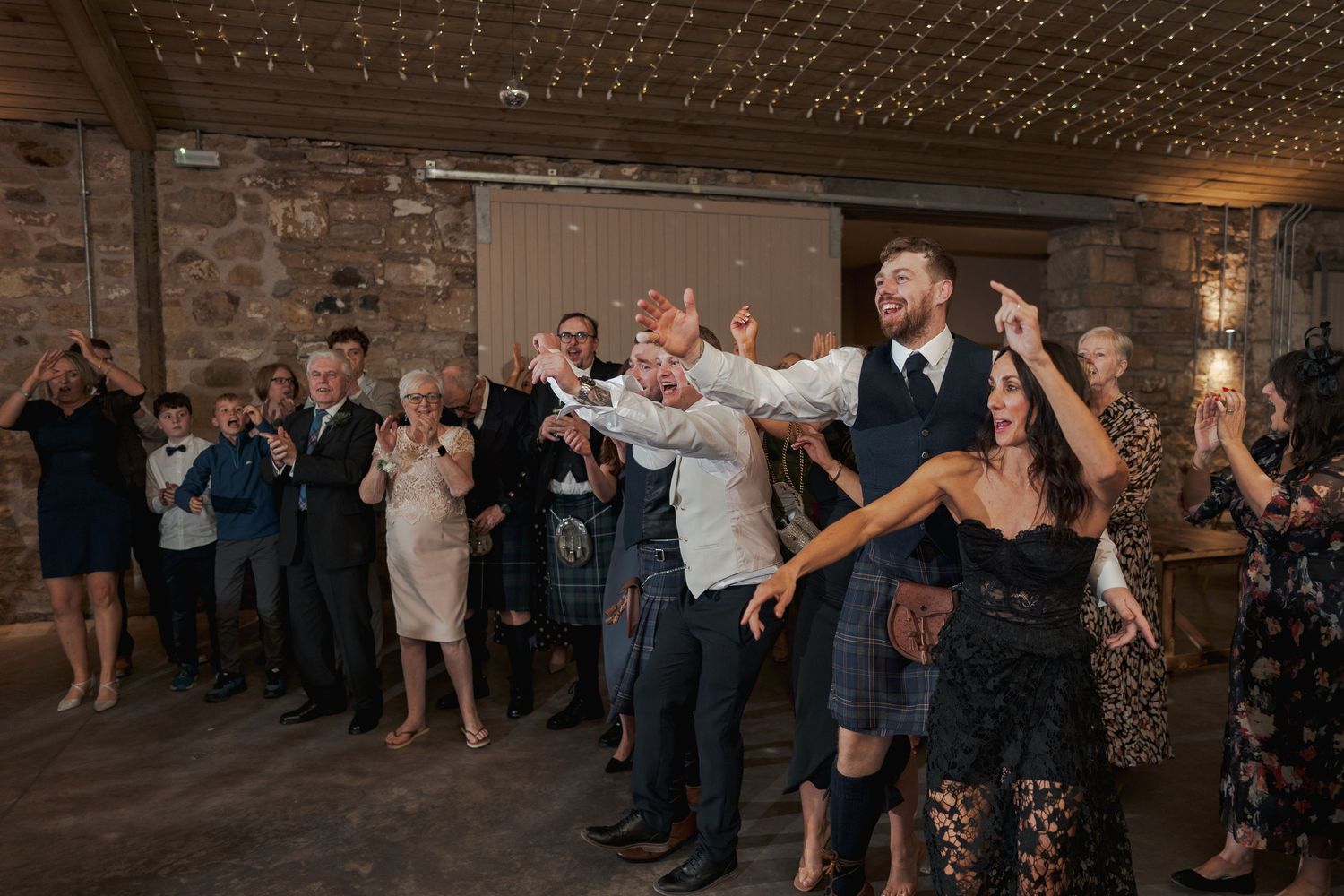Wedding guests dancing and celebrating together at Falside Mill on a rustic dance floor with exposed brick walls and string lights.