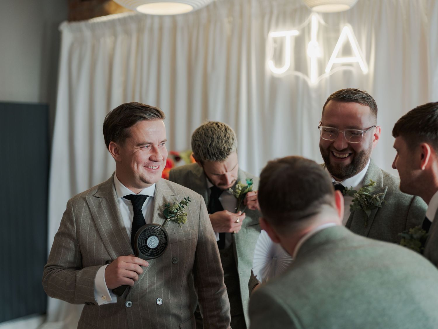 Wedding guests in formal attire gathered at a reception venue with white curtain backdrop.