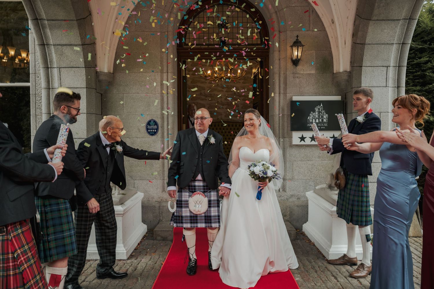 Bride and groom in Scottish kilt exit venue on red carpet as guests throw colorful confetti outdoors.