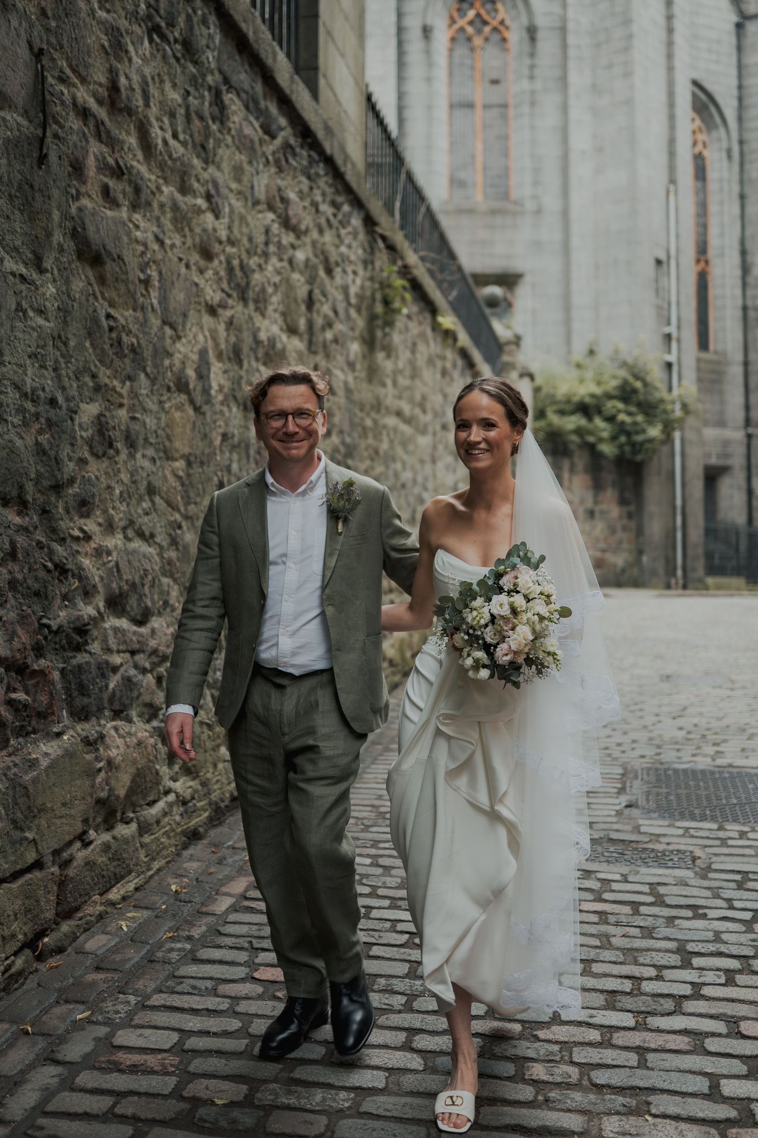 Bride and groom walk hand-in-hand on cobblestone street beside stone church, she holds pastel bouquet.