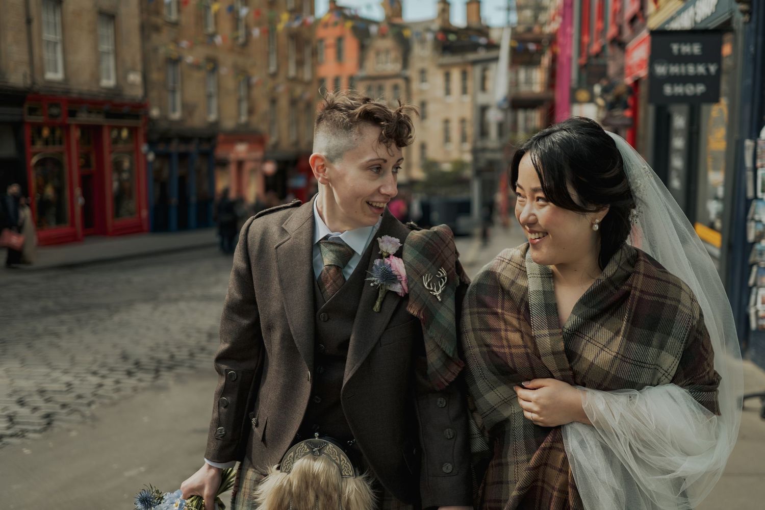 Newlywed couple smiling on a cobblestone street in Edinburgh, bride in veil and groom in tweed suit.