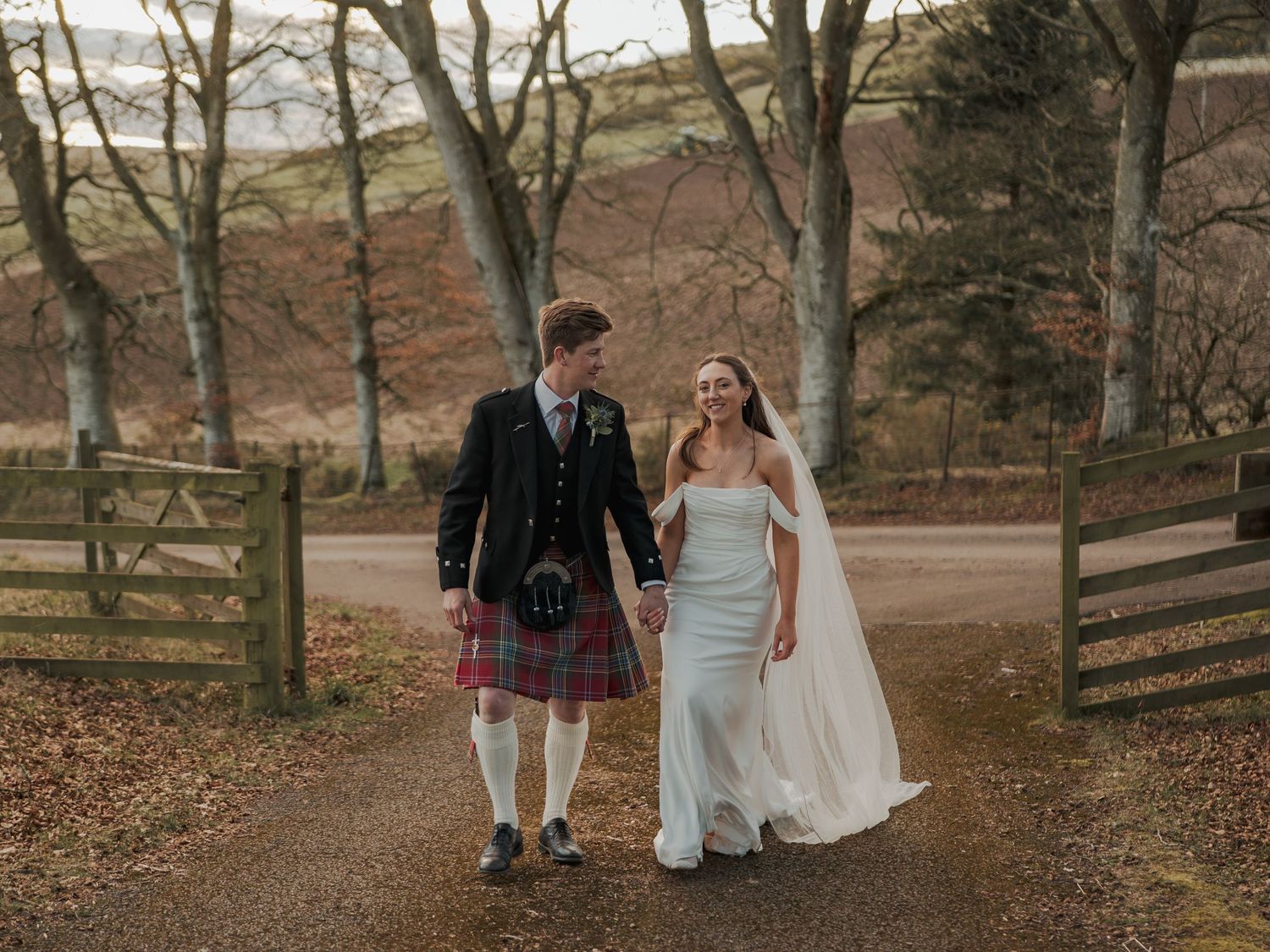 A bride and groom walk hand in hand on a Scottish estate, groom wearing a tartan kilt, surrounded by autumn trees at Kinclune House