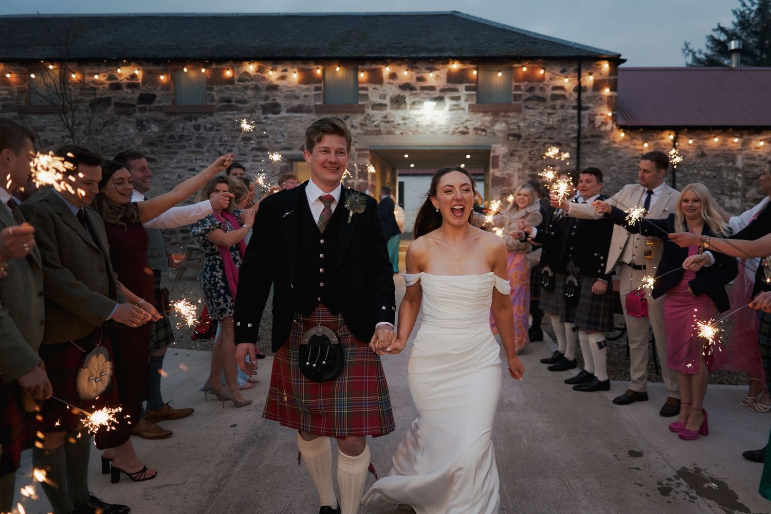 Newlyweds exit through sparkler tunnel at Scottish barn wedding, groom in traditional tartan kilt.