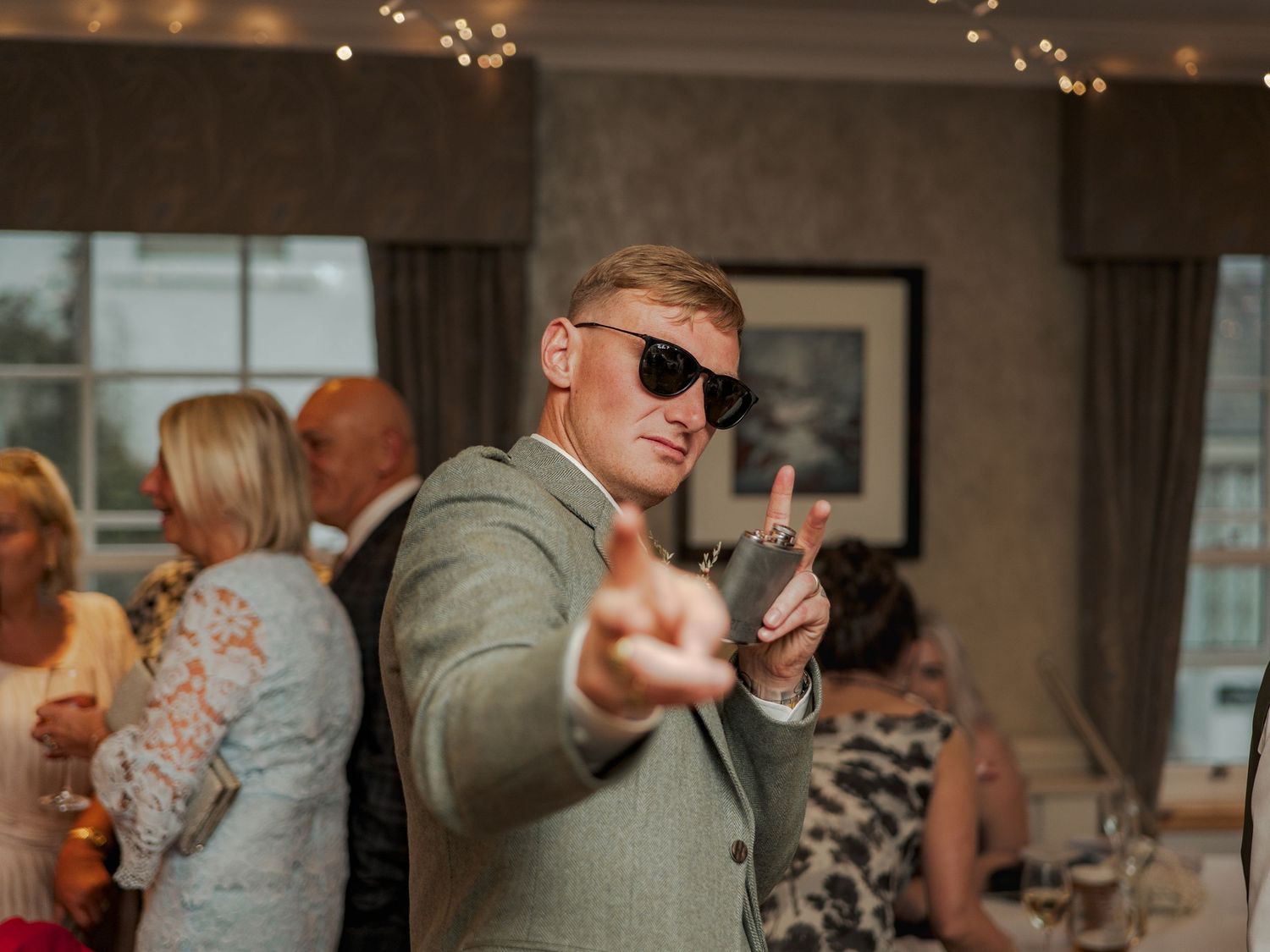 Guest gestures peace sign while wearing sunglasses and olive jacket at indoor celebration event.