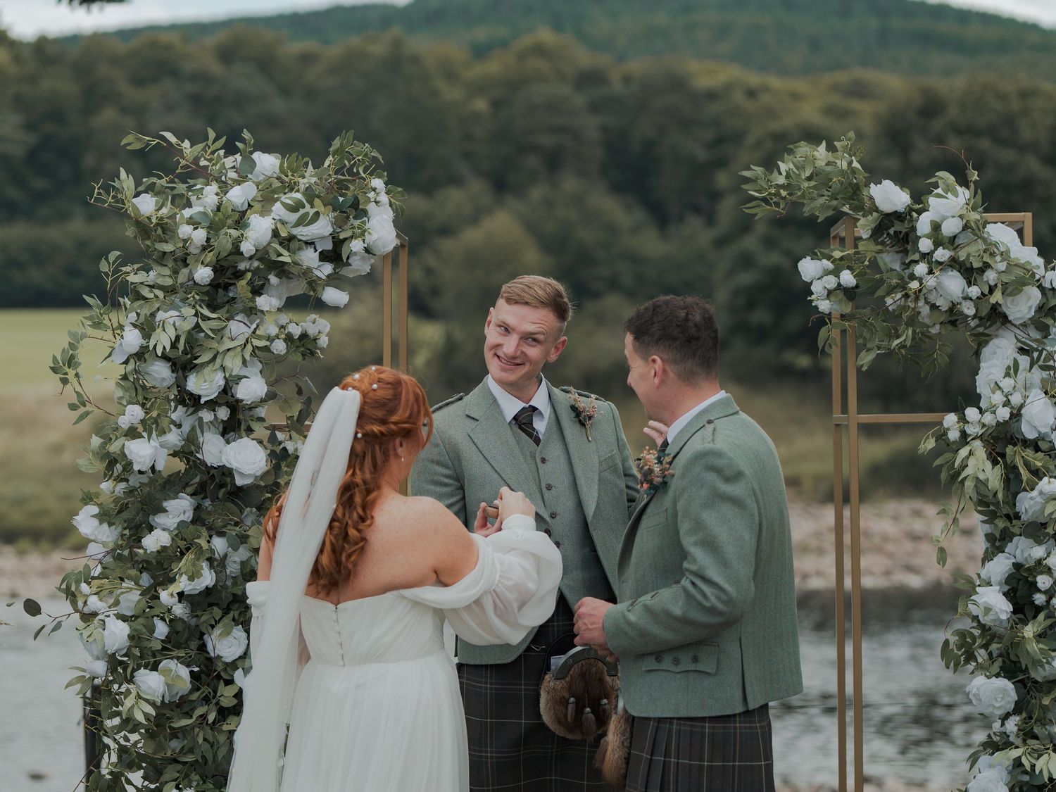 Wedding celebration moments captured by a lake with elegant white floral arrangements and pastoral scenery in the background at Banchory Lo.