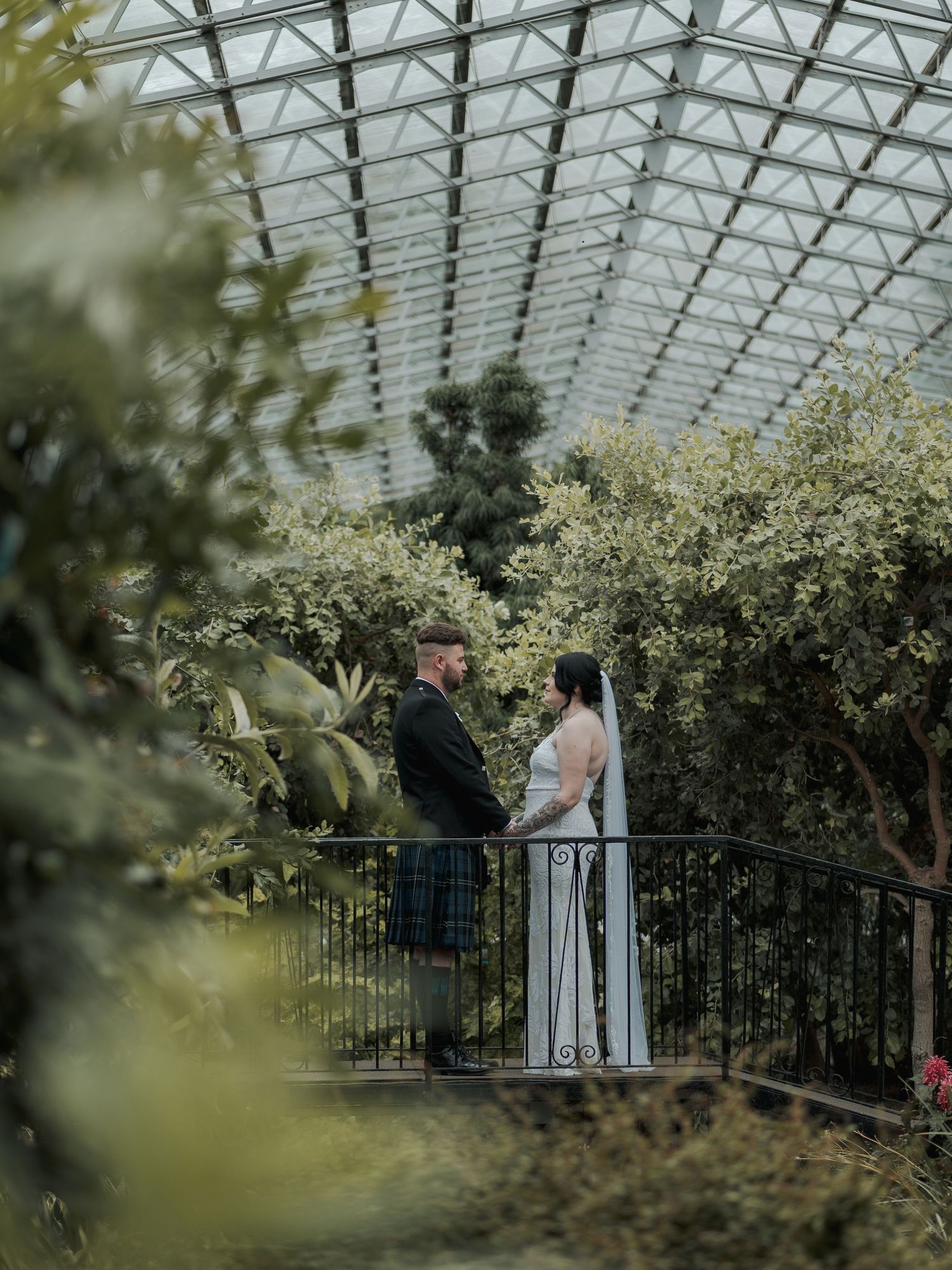Couple in wedding attire standing on a bridge inside a botanical garden greenhouse with lush greenery.