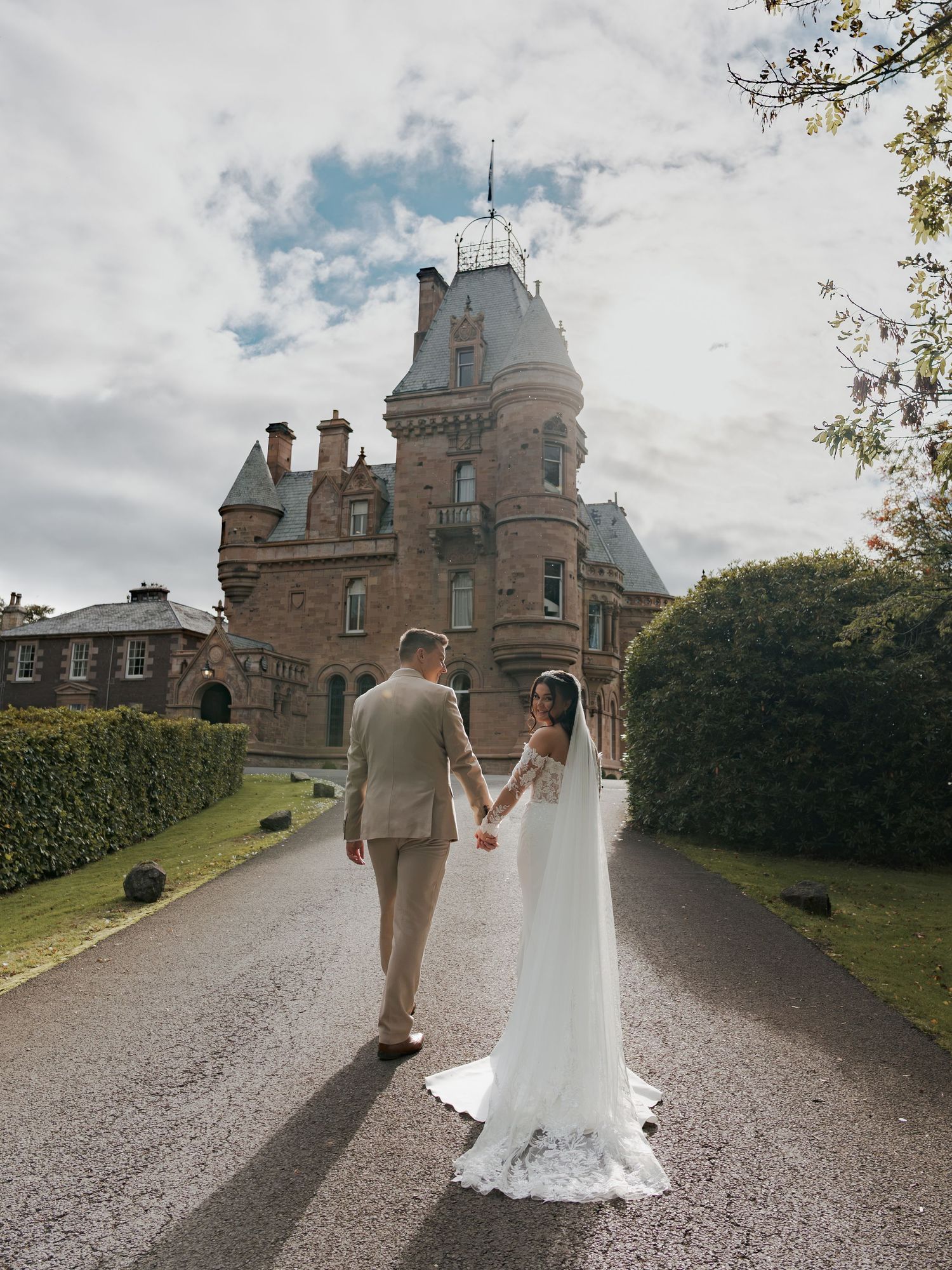 Bride and groom holding hands on a tree-lined path before a grand Scottish castle on their wedding day.
