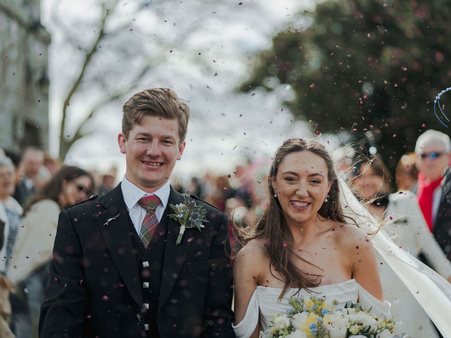 Happy newlyweds walking through confetti shower after wedding ceremony, bride in white gown holding bouquet, groom in tartan suit.