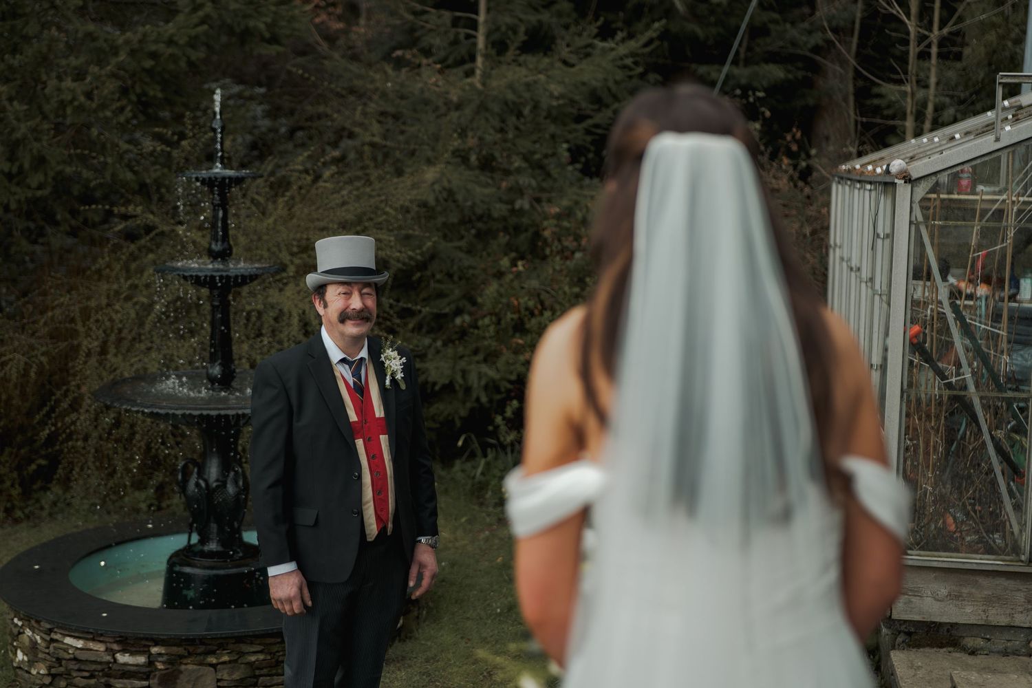 A smiling groom in a gray top hat and red tie reacts to seeing his bride in a white wedding dress outdoors.