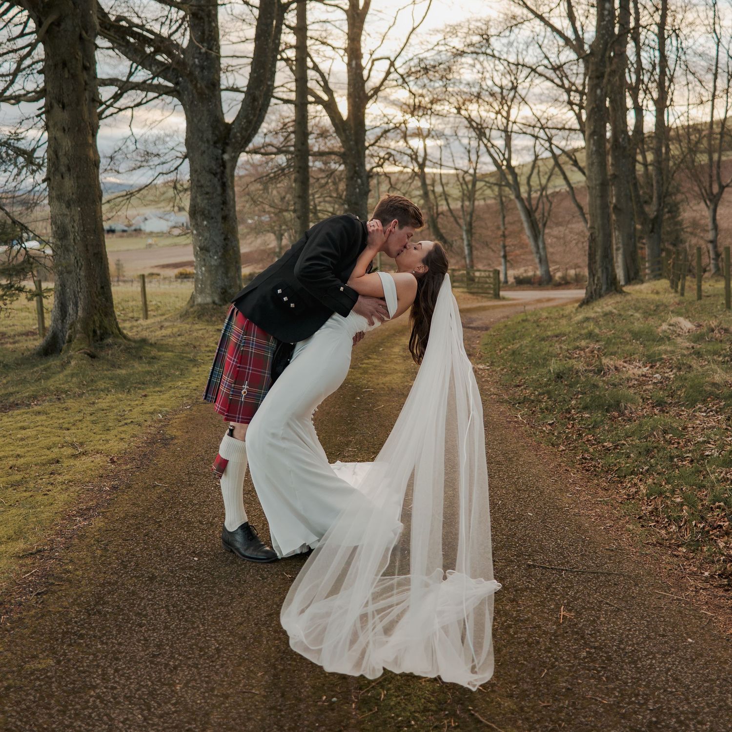 Bride and groom share a romantic dip kiss on a woodland path, groom in Scottish tartan kilt, bride in elegant gown with veil.