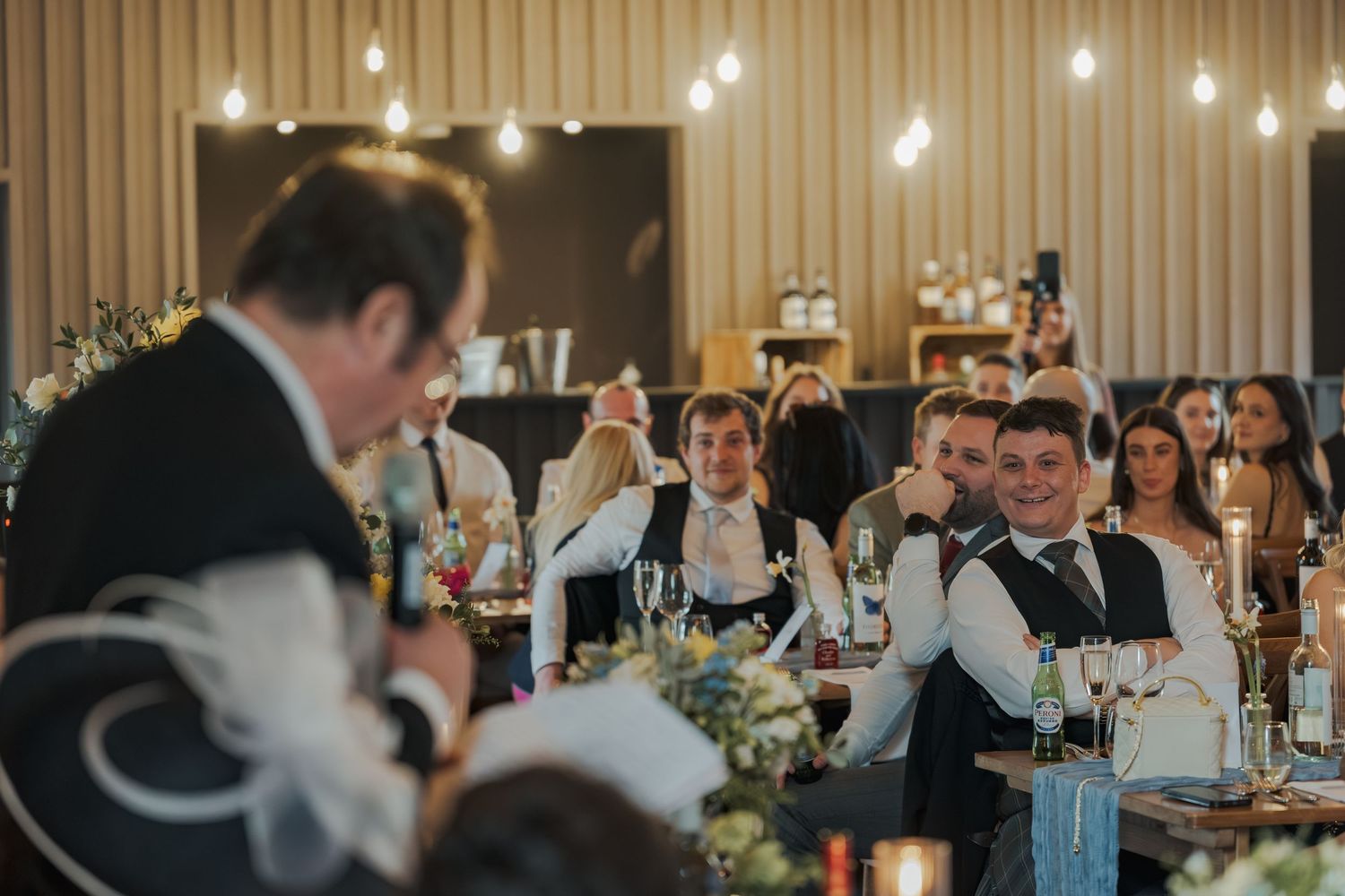 Guests laughing and raising glasses during wedding reception speeches in an elegantly lit venue.
