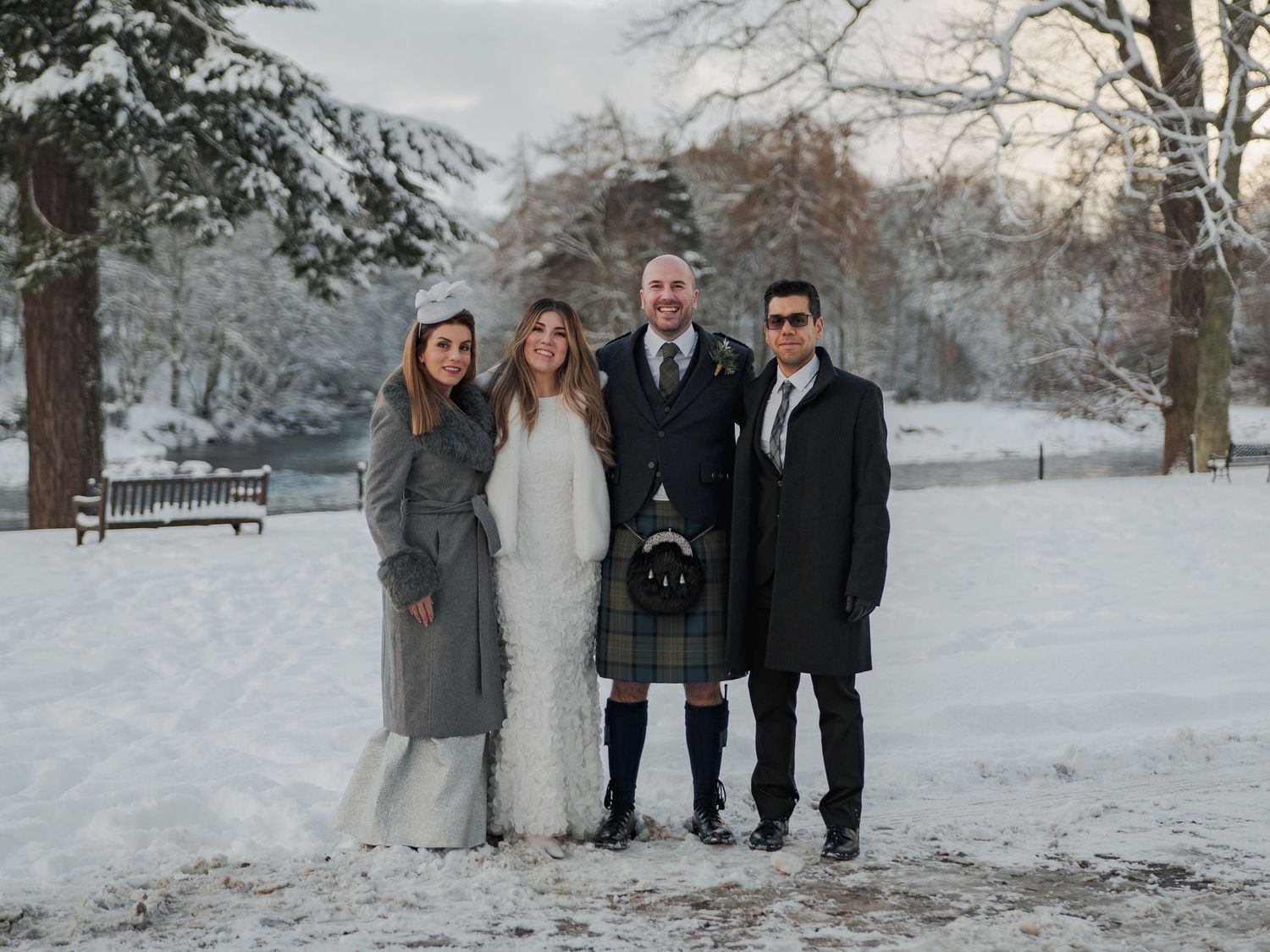 Four people posing in a snowy park, one man wearing a kilt, celebrating a winter wedding in Scotland.