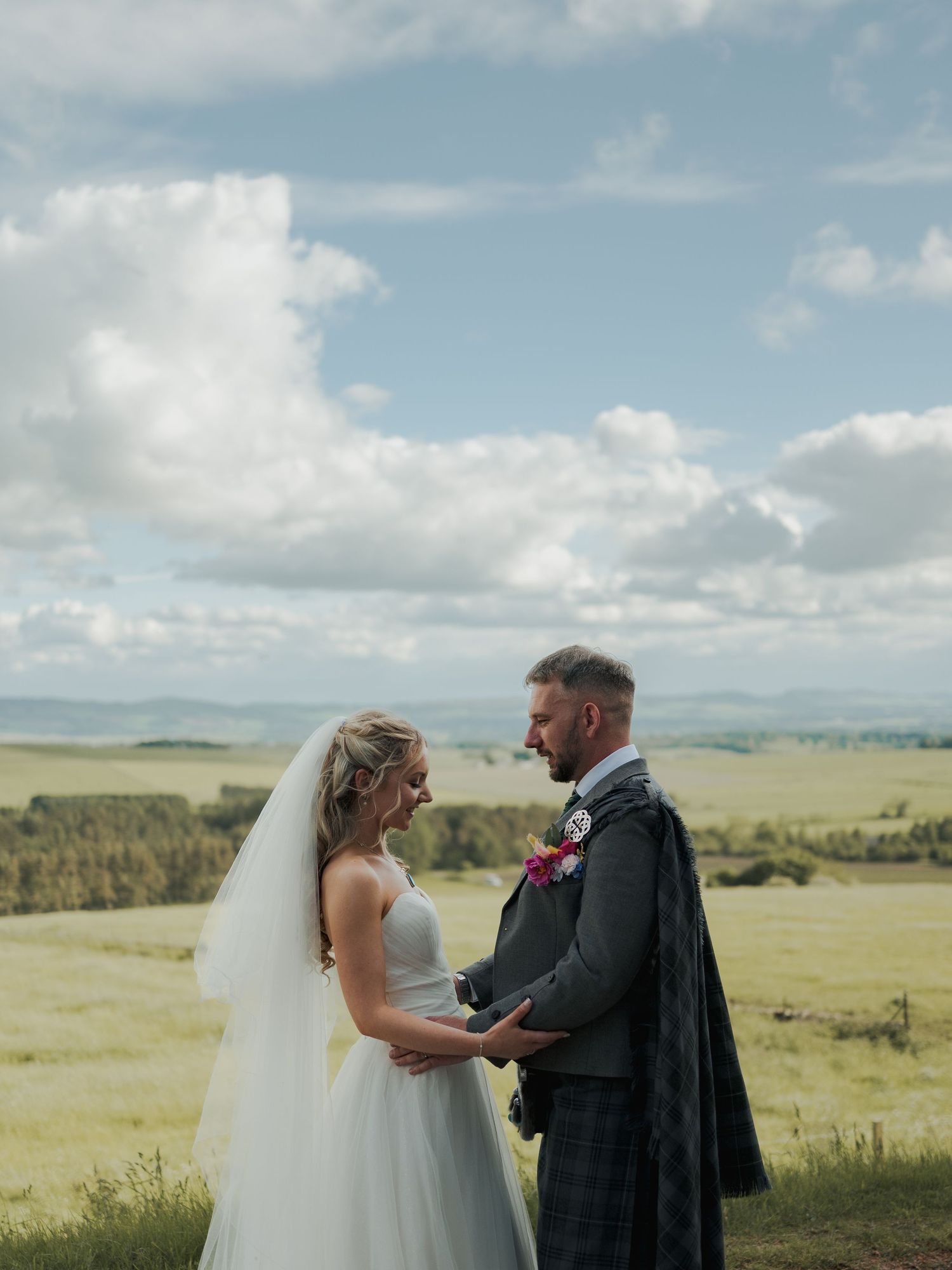 Bride and groom holding hands outdoors with scenic Scottish countryside and cloudy sky backdrop.
