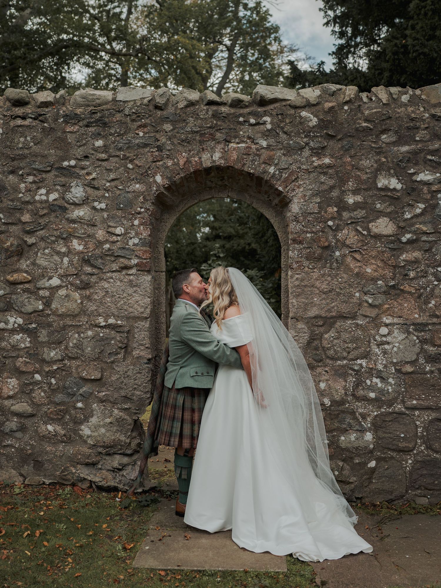 Bride and groom kissing in a rustic stone archway, groom wearing a Scottish kilt, bride in a white gown with veil.