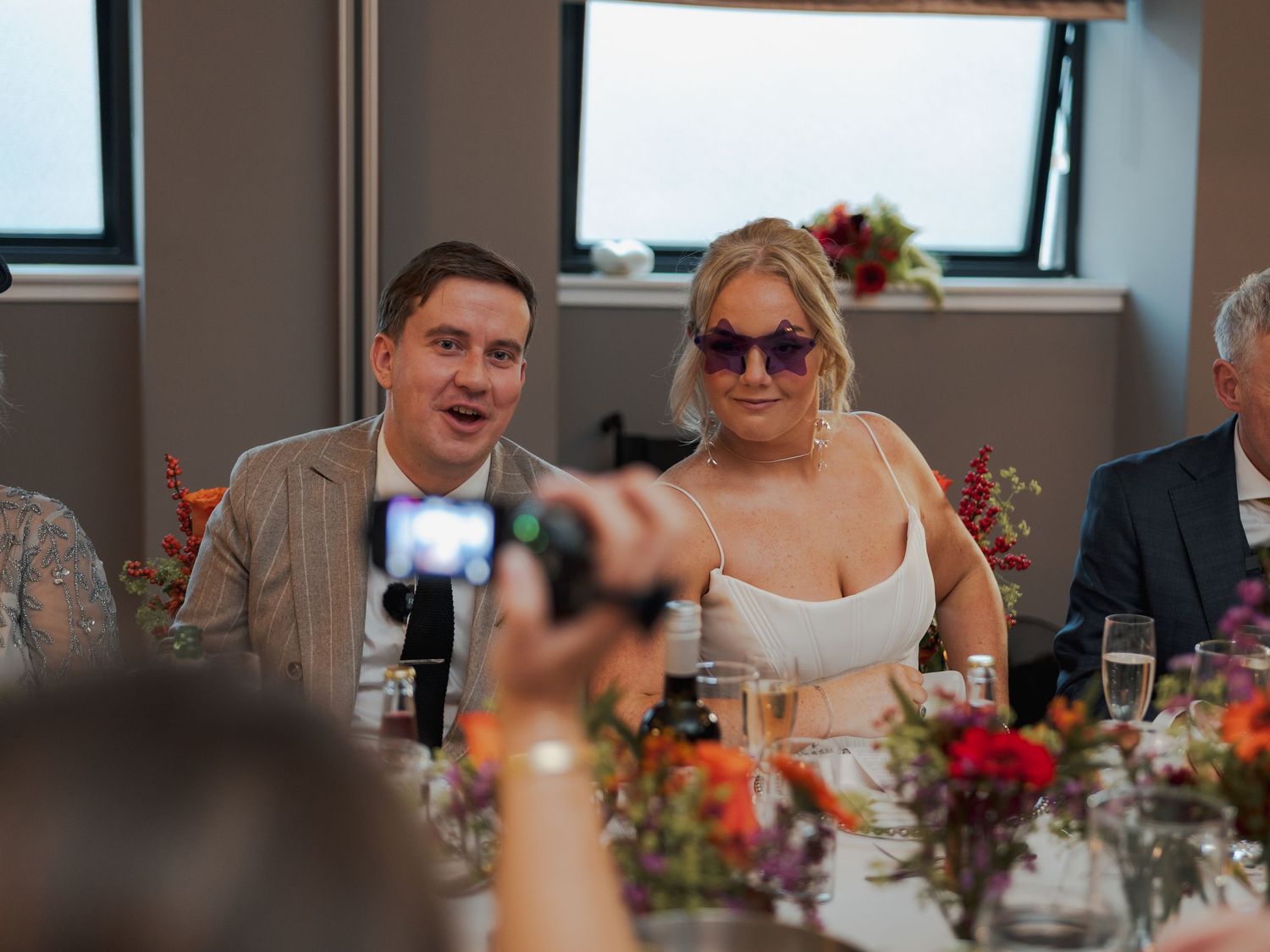 Wedding guests seated at tables decorated with colorful floral centerpieces during an indoor reception.