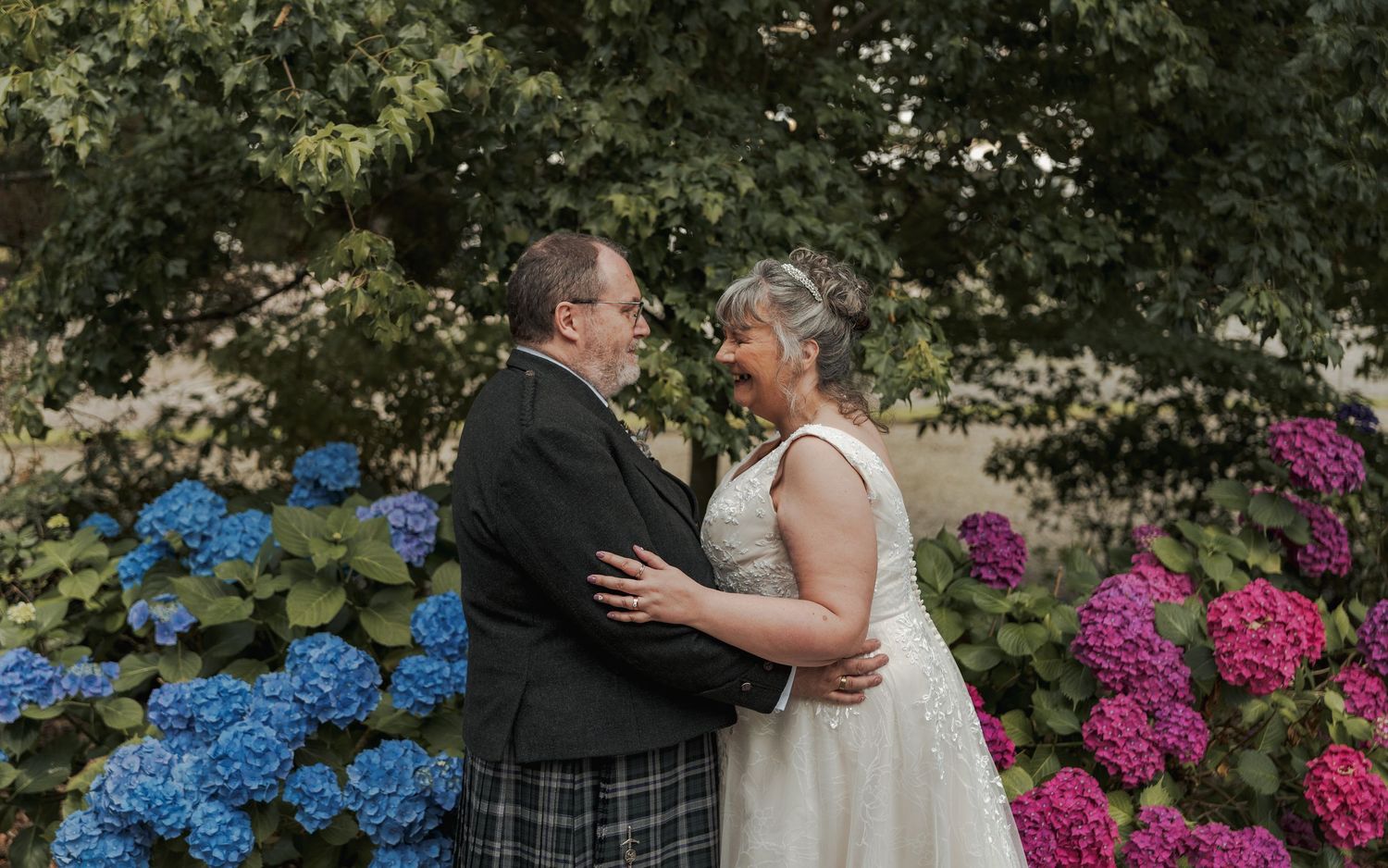 Couple in wedding attire embracing among colorful hydrangeas, man in tartan kilt and woman in white gown.